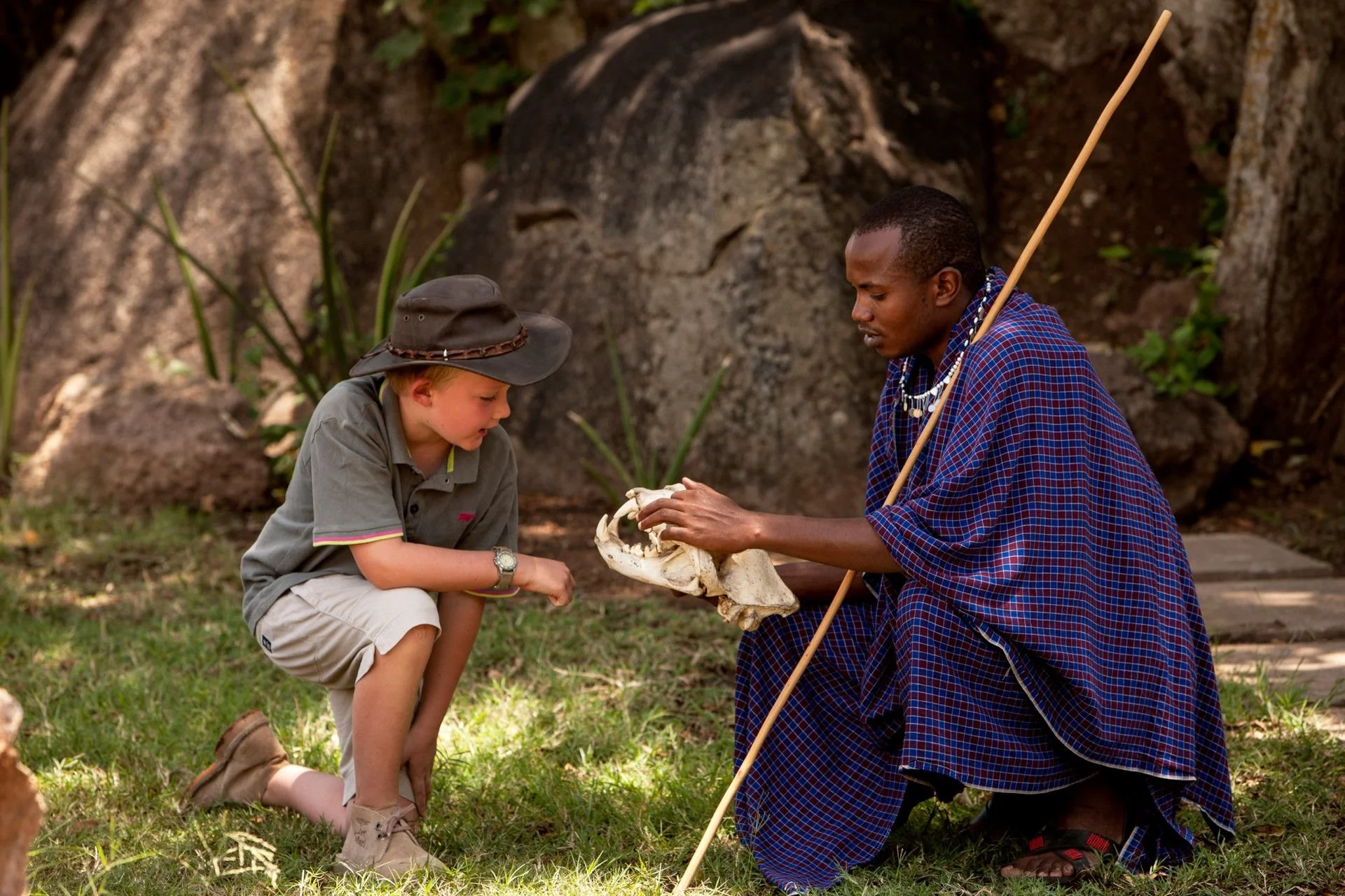 A man wearing traditional Maasai clothing and jewelry kneeling on the ground, holding a skull, and talking to a boy wearing a gray shirt, beige shorts, and a wide-brimmed hat, who is bent down looking at the skull.
