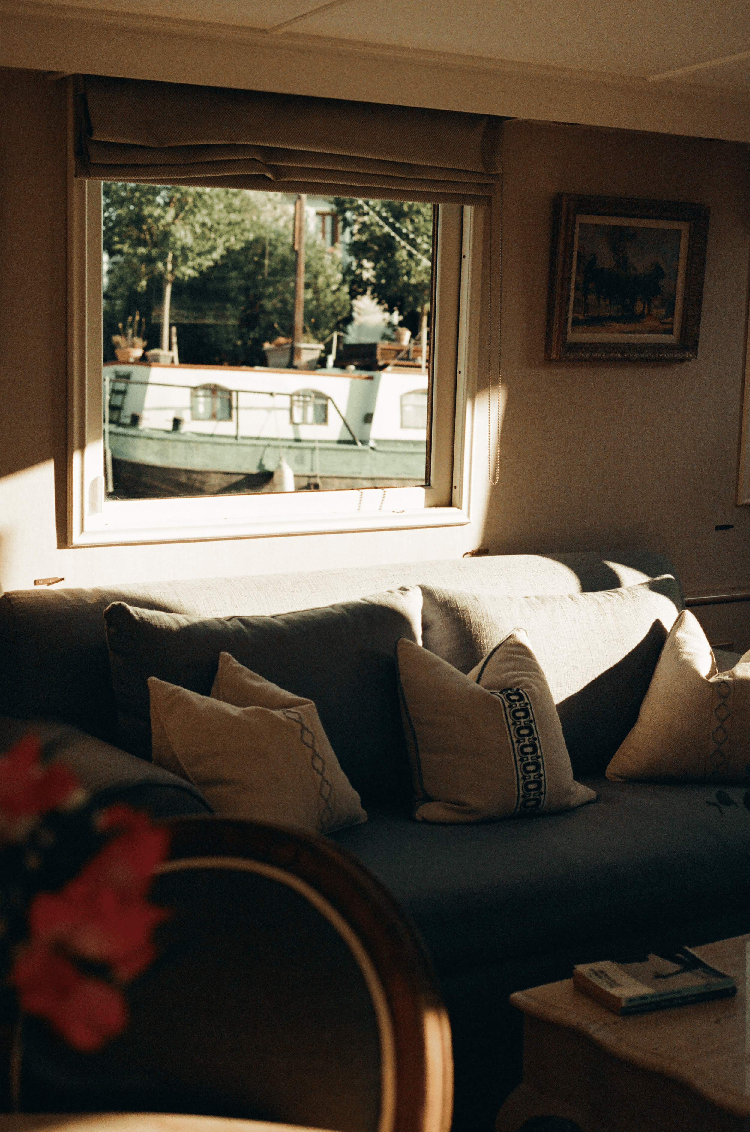 Living room with a window showing a boat and trees outside, beige sofa with patterned pillows, side table with a book, and framed artwork on the wall.