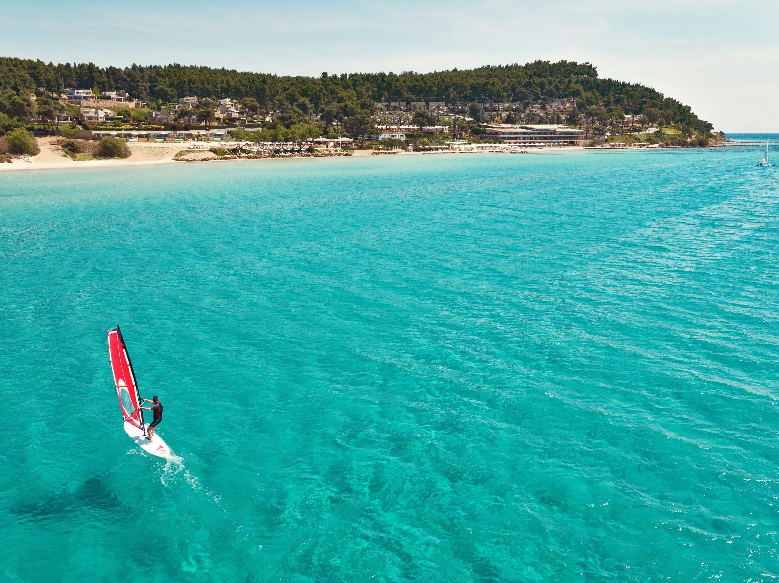 A person windsurfing on turquoise water near a seaside town with houses and green trees on a hillside.