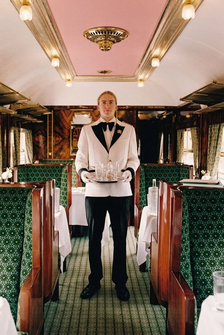 A waiter in a tuxedo holding a tray with glasses inside a vintage-style restaurant with green patterned booths and white tablecloths.