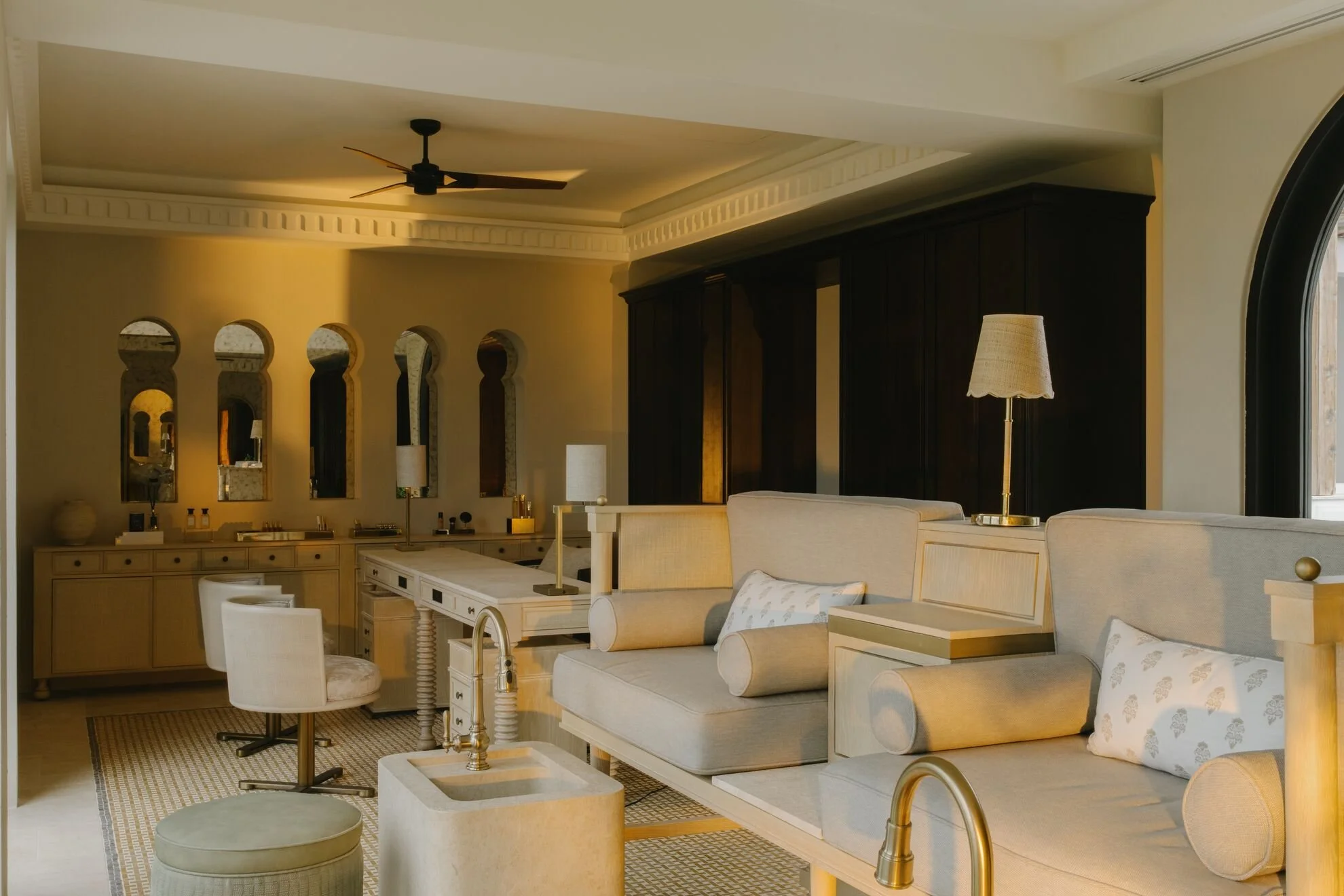 Spacious bedroom with beige sofas, decorative pillows, a cream-colored dresser, and a dark wood wardrobe. The room features a ceiling fan, an arched window, a standing lamp, and mirror-paneled walls with built-in lighting.
