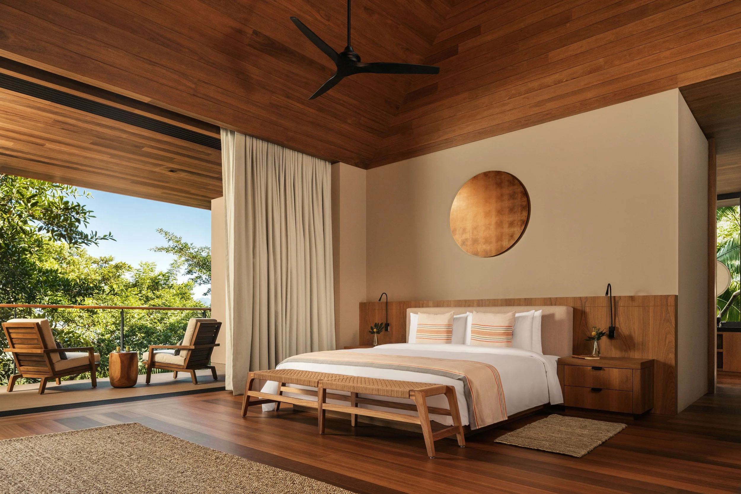 Modern bedroom with a large bed, wooden headboard, small side tables, and ceiling fan, with an outdoor balcony featuring chairs and green trees.