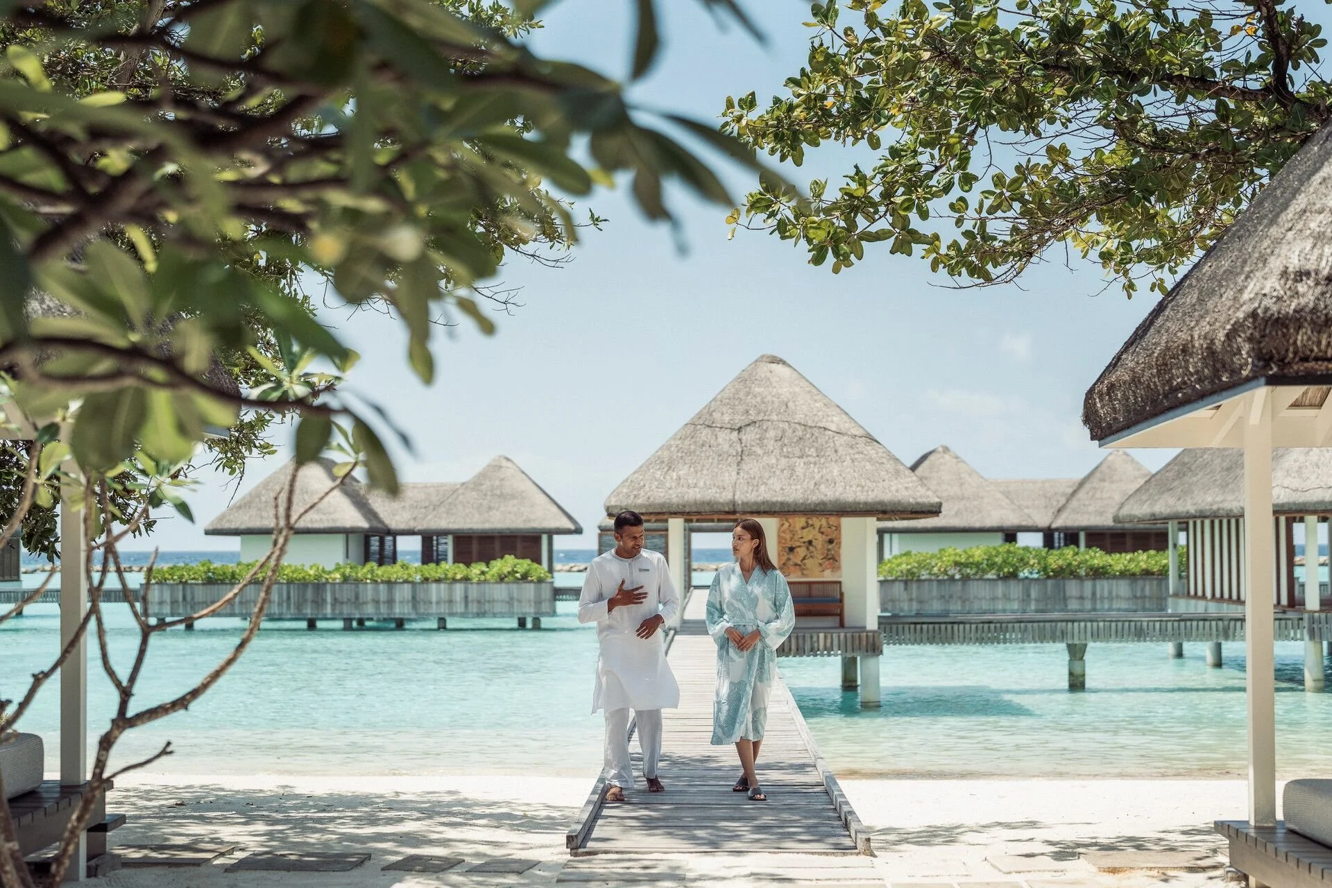 Two people, a man and a woman, walking on a wooden dock over water at a tropical overwater resort, with thatched-roof bungalows in the background, trees in the foreground, and a blue sky.