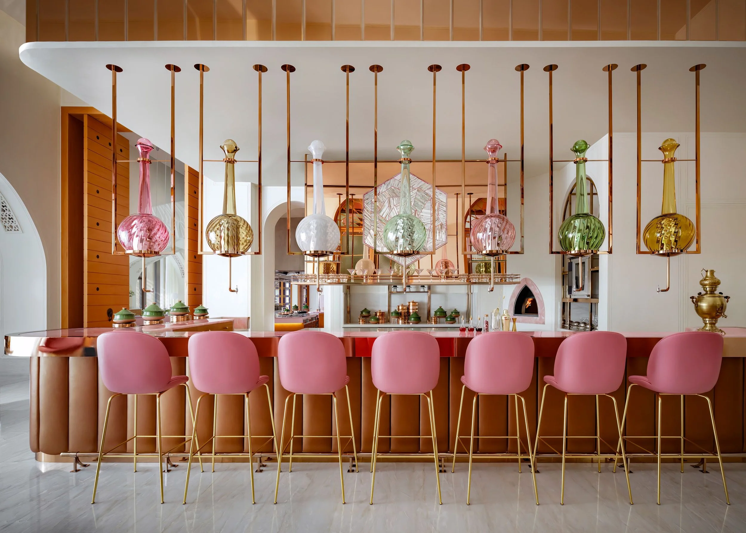 A modern dining area with pink chairs around a round table, decorative glass bottles hanging from a copper metal frame, and an interior design with wooden and white accents.
