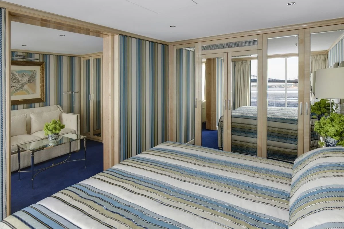 A bedroom with striped bedding, a mirrored closet, a sitting area with a white sofa, a glass coffee table, and room with large windows showing an outside view.