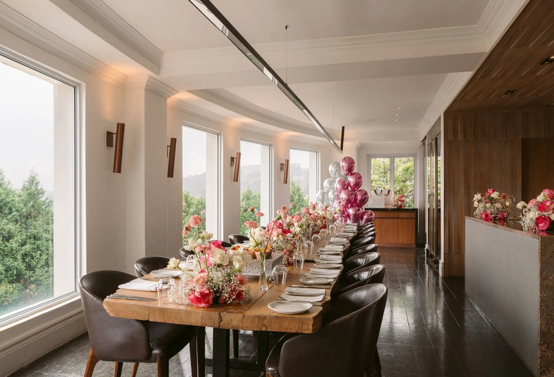 A decorated dining room for a celebration with a long wooden table, floral centerpieces, pink and white balloons, and natural light coming through large windows.