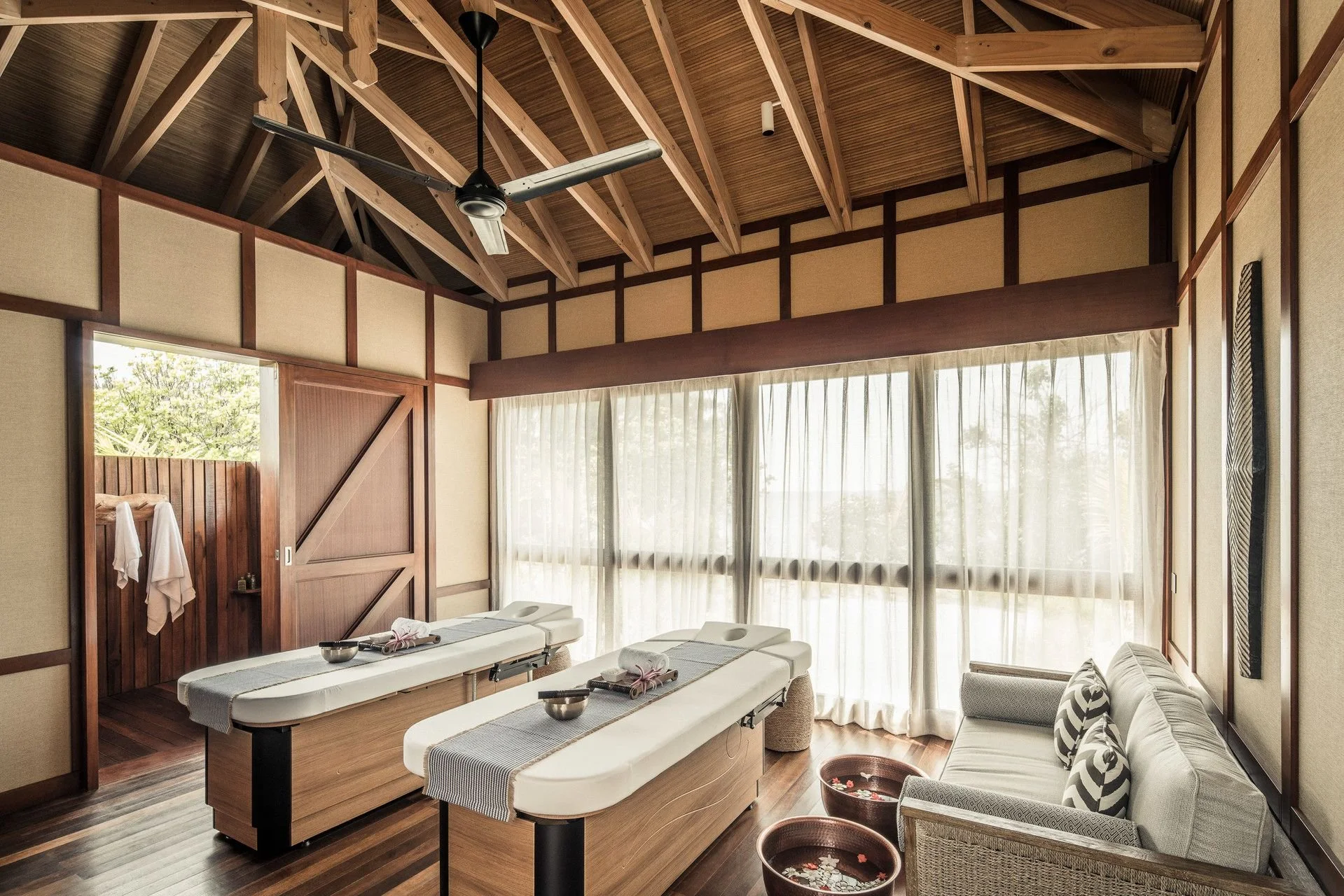 A bright interior room with wooden beams, large windows with curtains, and massage tables with towels, in a spa or massage therapy setting.
