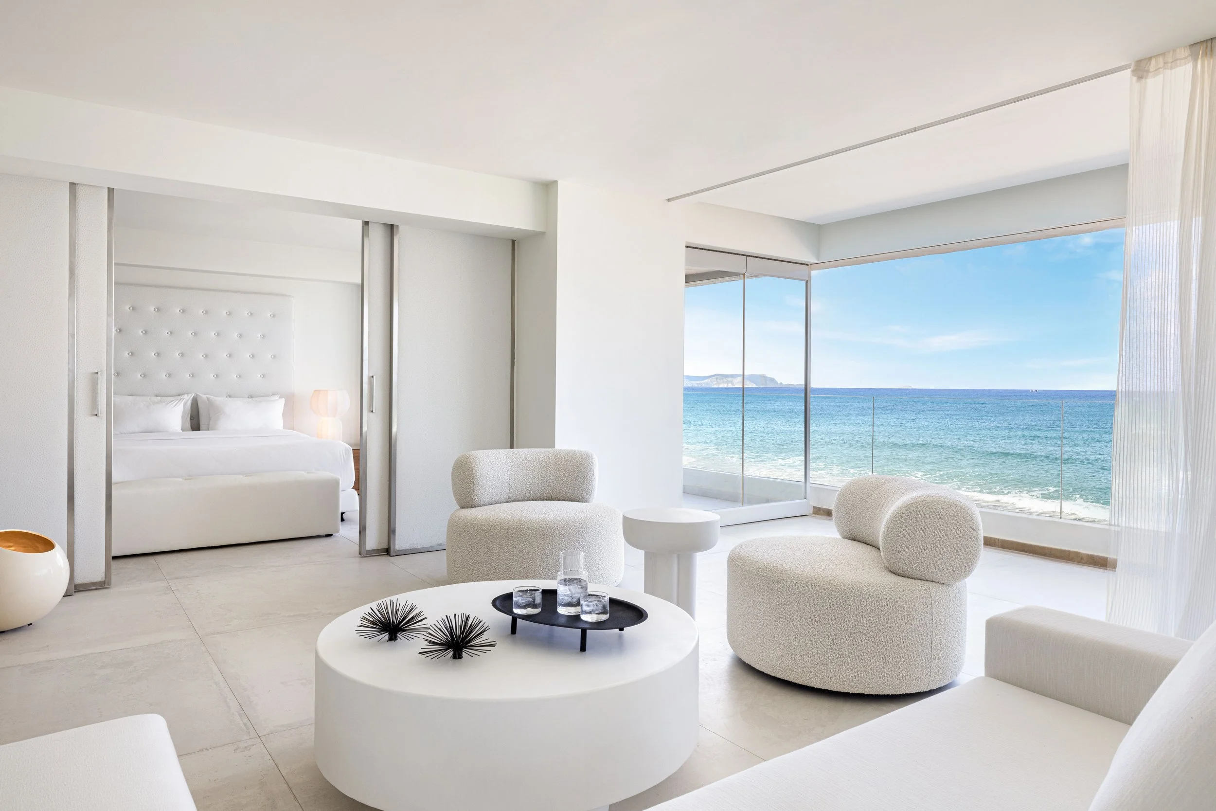 A modern, white living room with large windows showing an ocean view. There are white chairs, a white bed, and minimalist decor, including black accent pieces on a round coffee table.