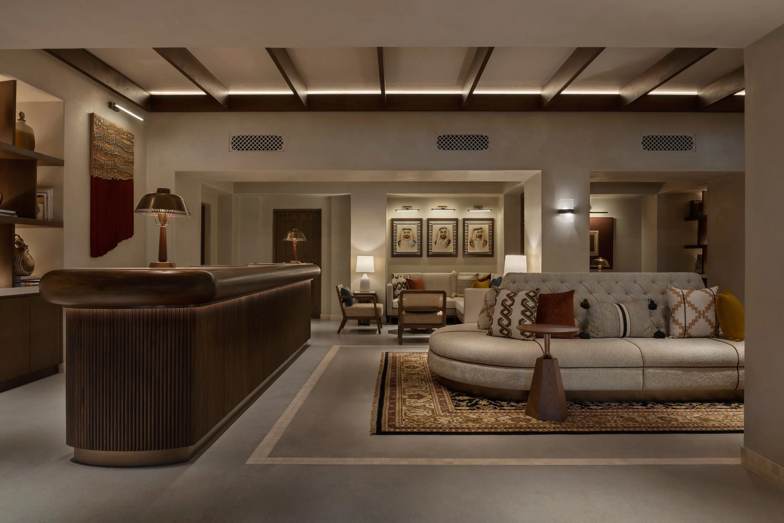 A spacious, well-lit living room with modern decor featuring framed portraits on the wall, a curved sectional sofa with patterned pillows, a traditional area rug, and a sleek wooden bar counter.