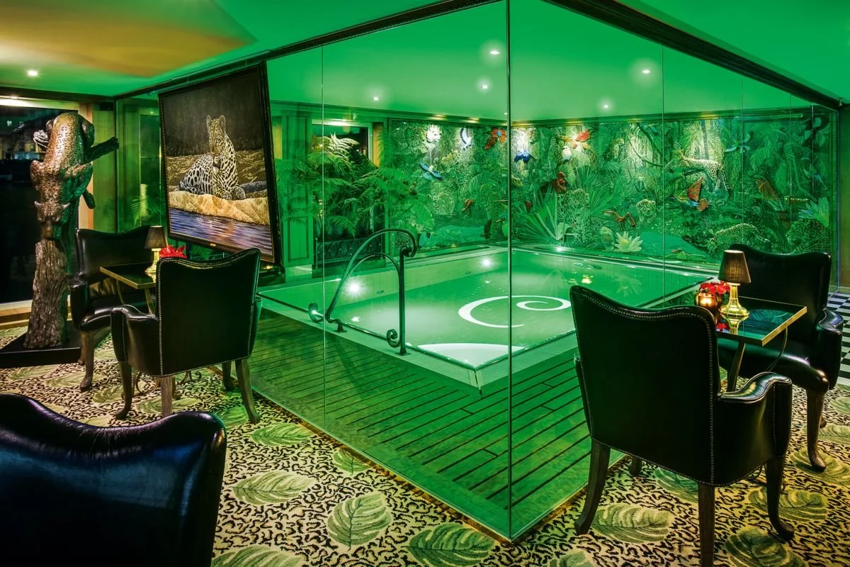Interior of a lounge with a small pool, green lighting, lush jungle wallpaper, black leather chairs, and a large painting of a leopard.