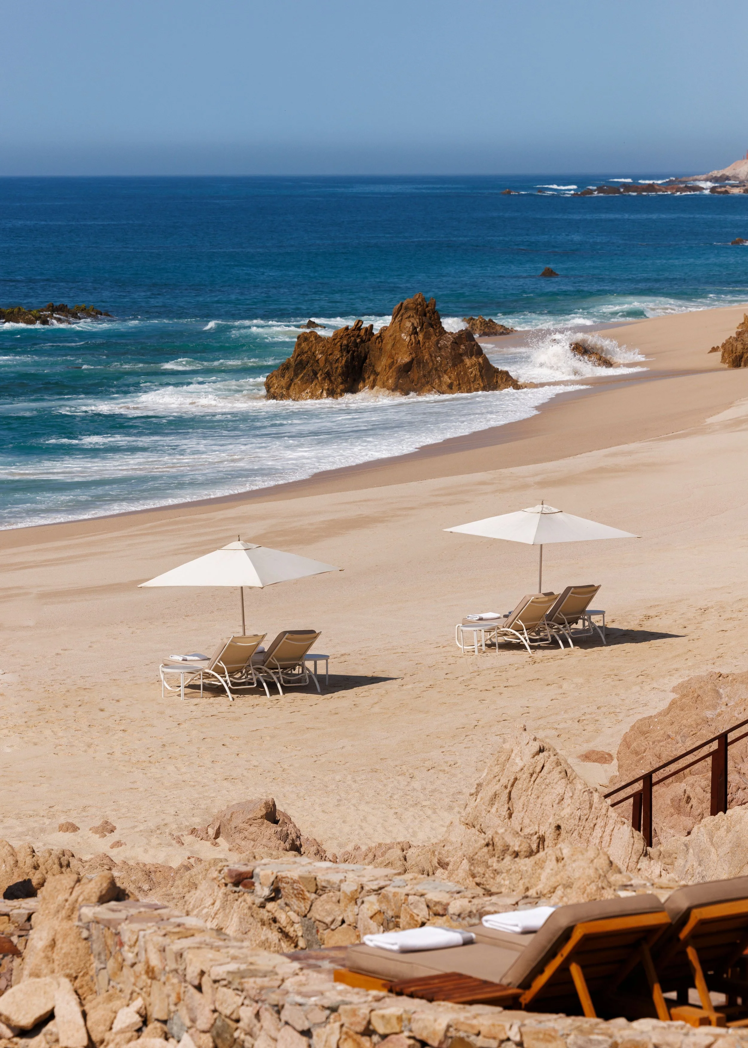 A serene beach scene with two lounge chairs and umbrellas on sandy beach, rocky shoreline, and ocean waves crashing on rocks with a clear blue sky.