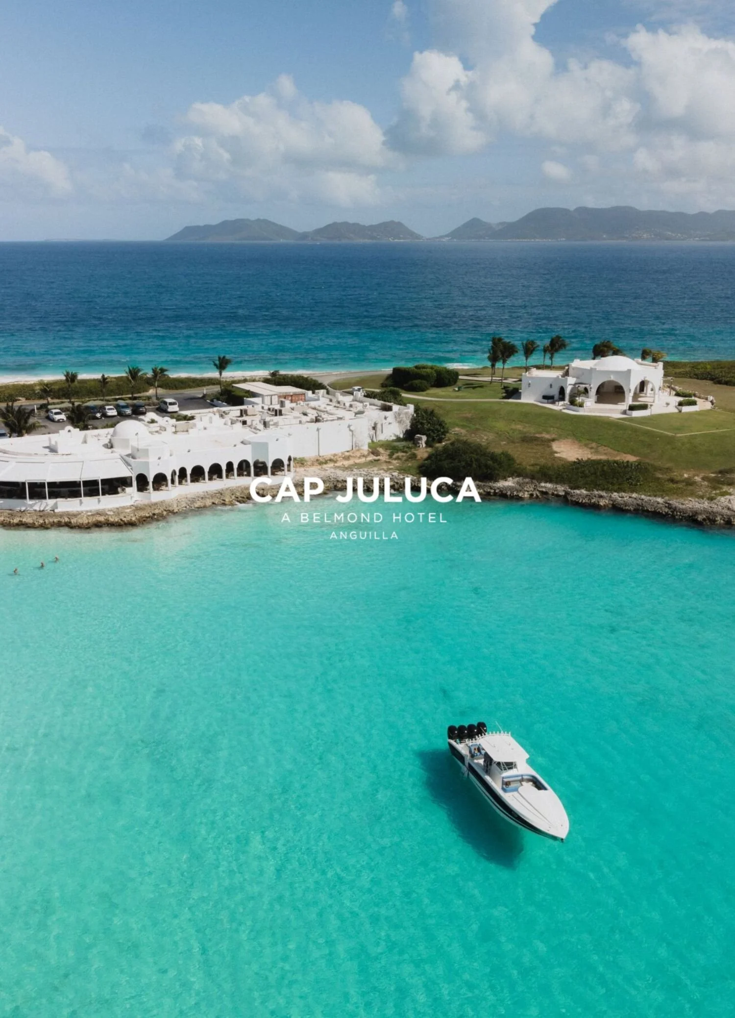 Aerial view of turquoise water with a white yacht and white buildings on the shore in Cap Juluca, Anguilla, with palm trees and green landscape, mountains in the background, and partly cloudy sky.