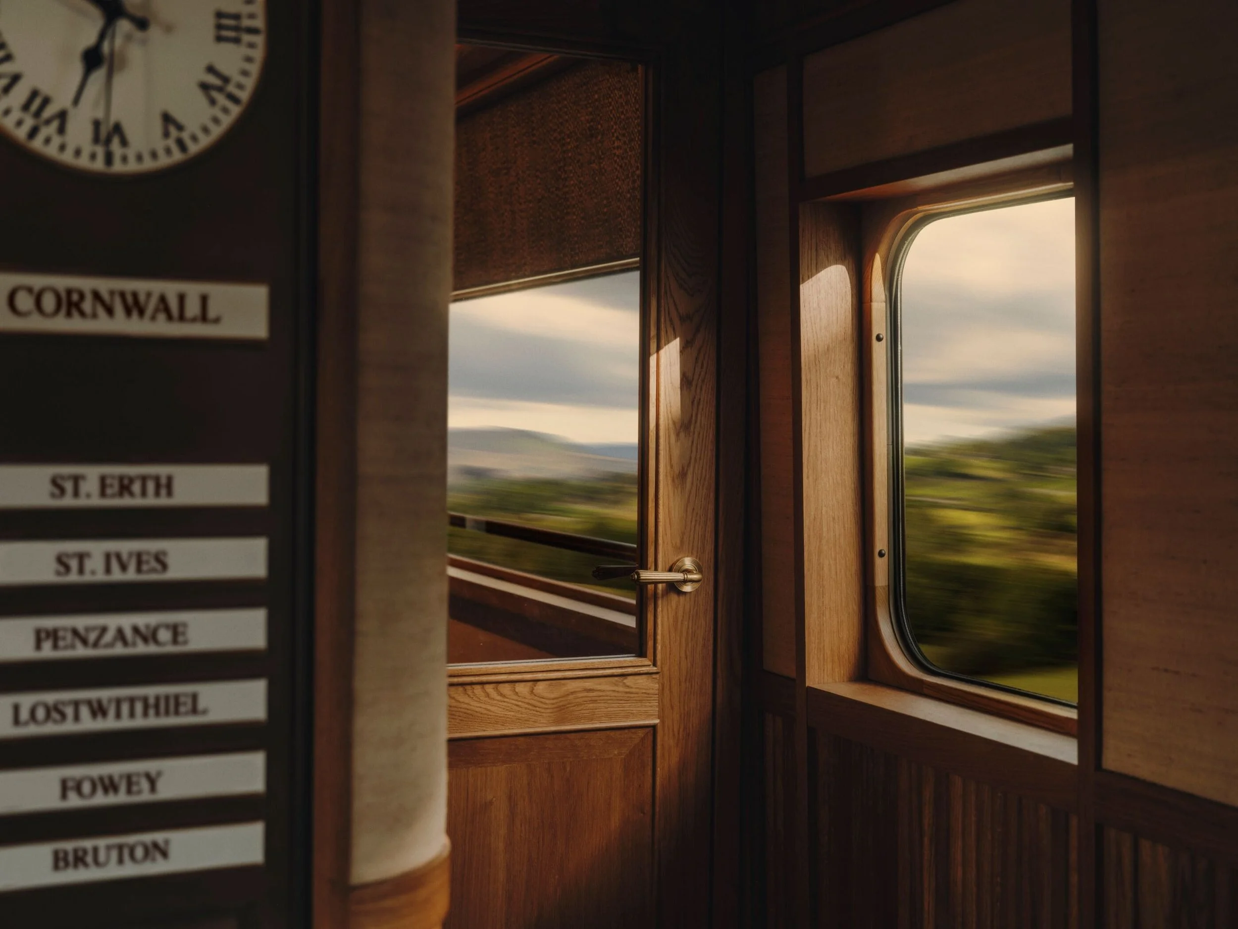 Inside a wooden train car, looking out the window at countryside with green fields and a cloudy sky. A station sign with station names is visible on the left side.