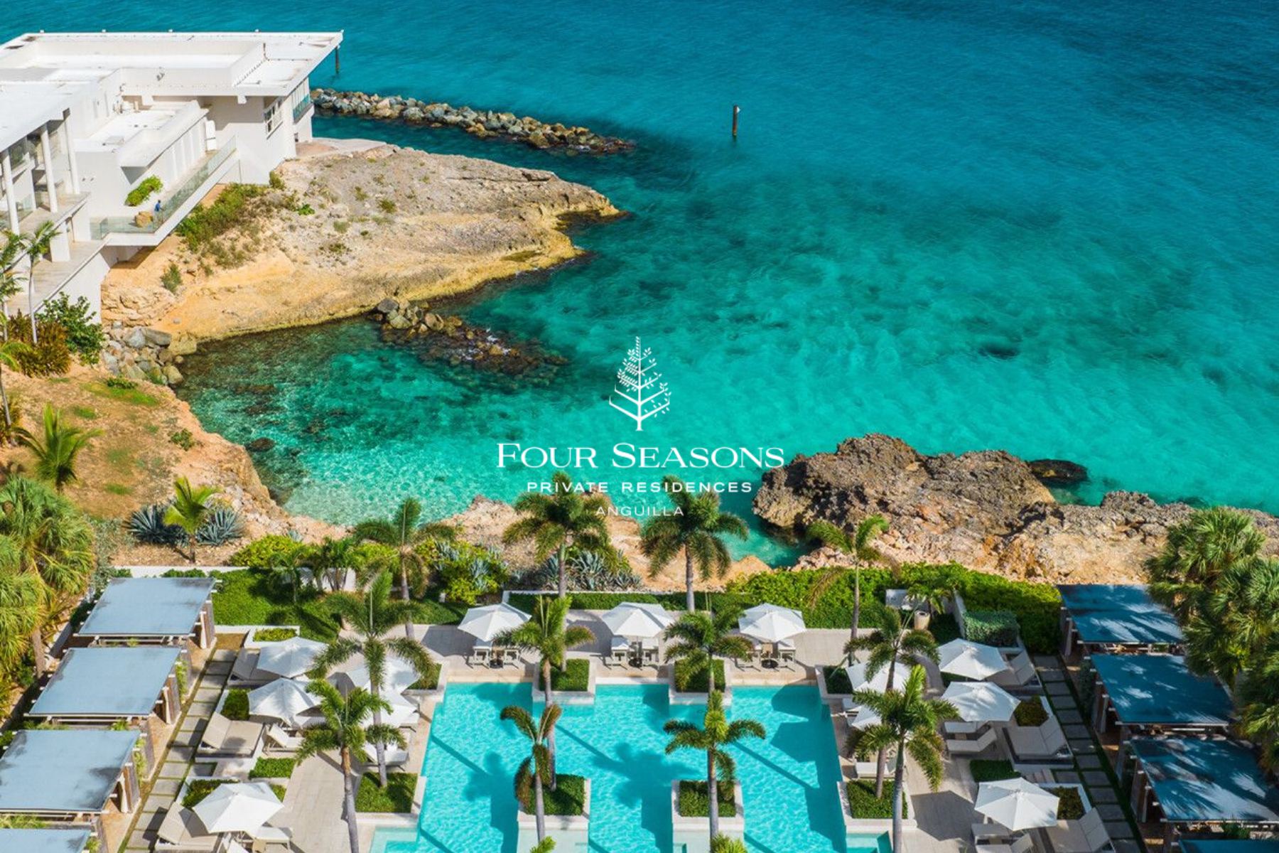 Aerial view of a luxury oceanfront resort with swimming pools, palm trees, white umbrellas, and lounge chairs, adjacent to turquoise ocean water and rocky coastline in Anguilla.