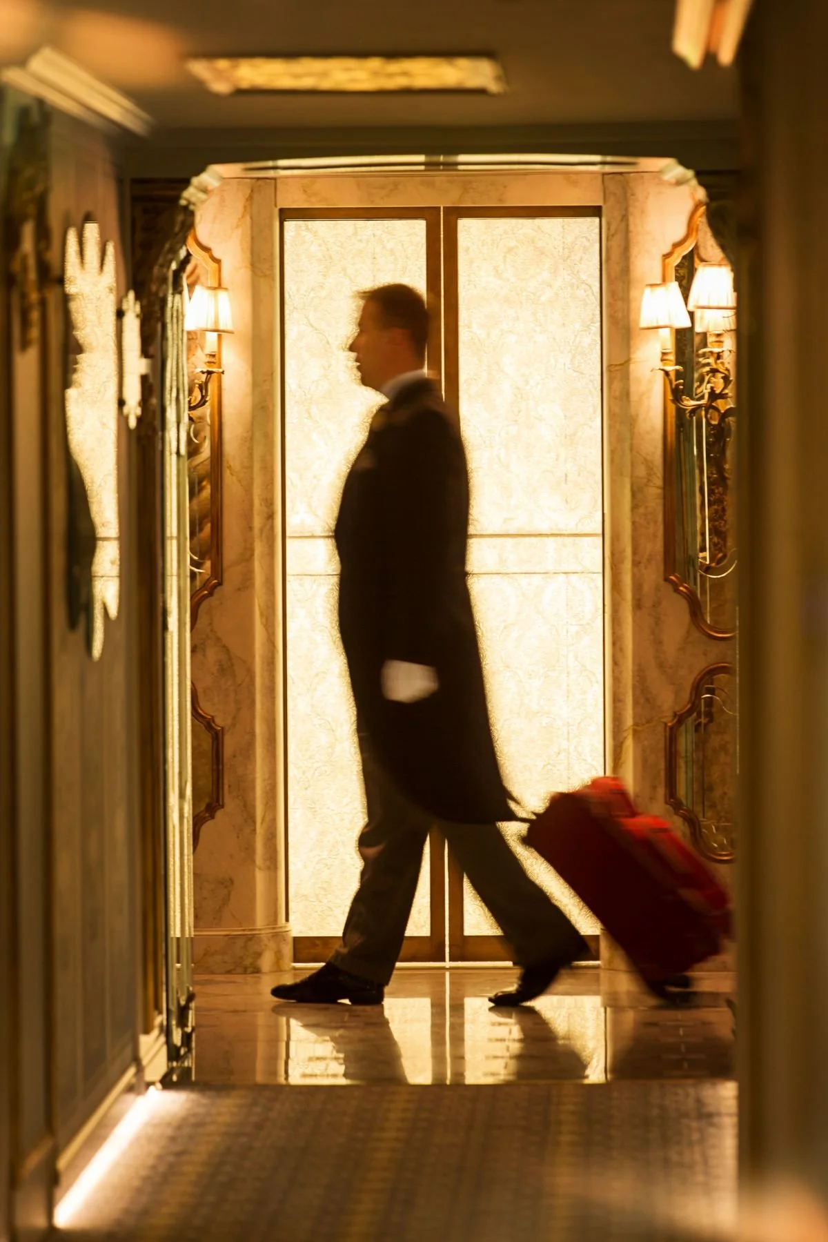 A person walking with a rolling suitcase through a hotel corridor, with a backlit door in the background.