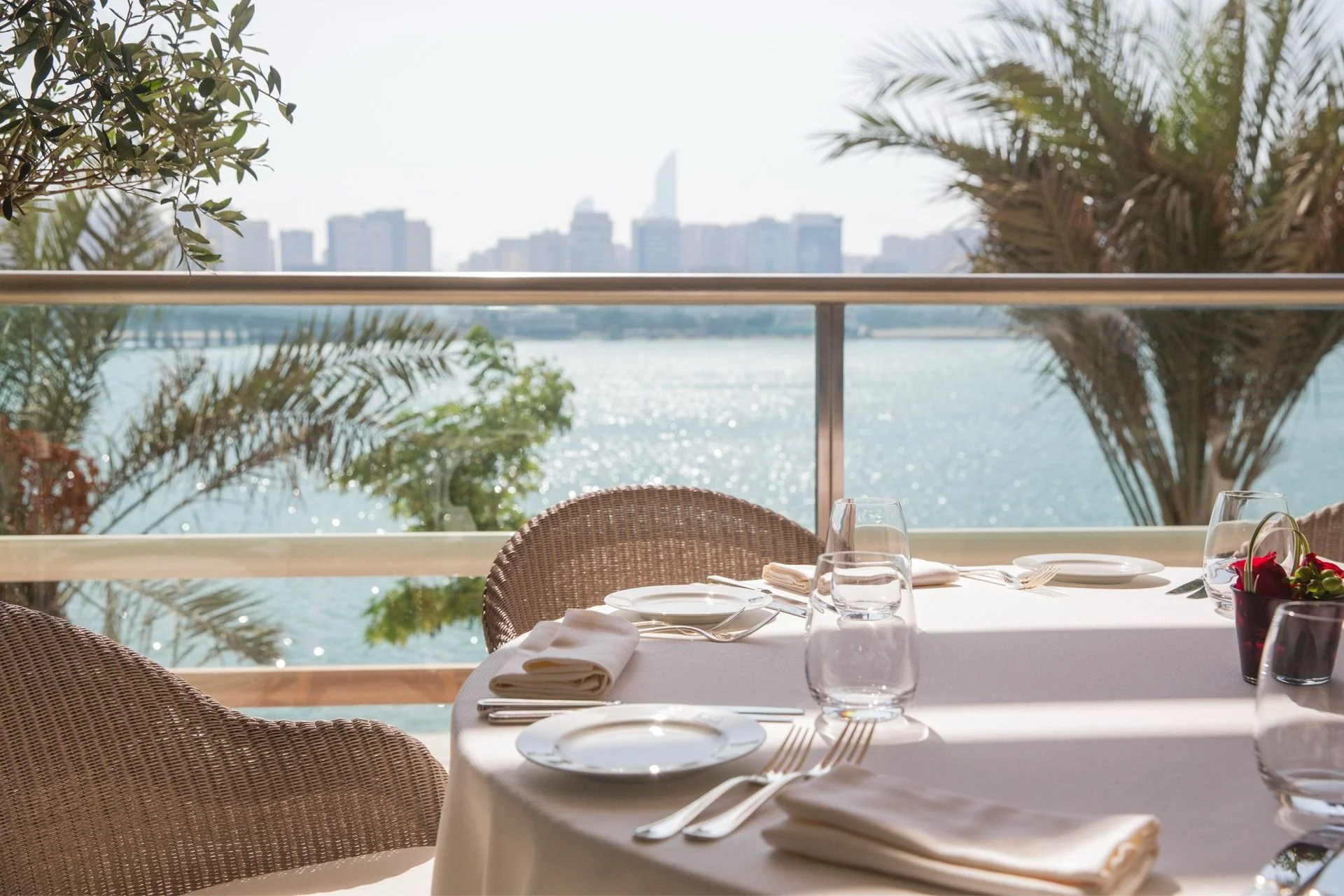 Elegant dining table set by a large window with a view of water and city skyline in the background, featuring white tablecloth, glassware, plates, silverware, and floral centerpiece.