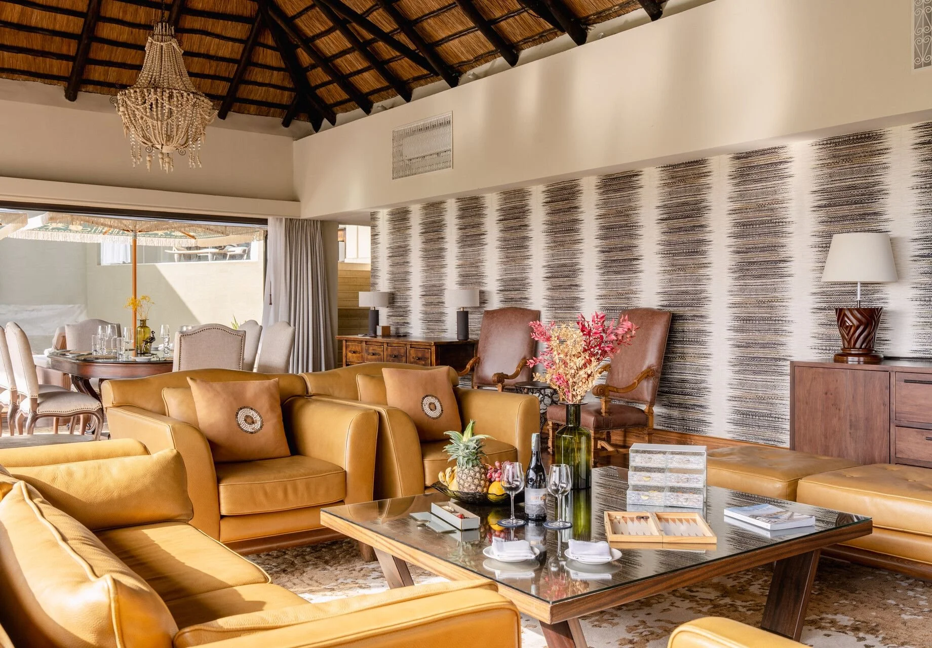 A spacious living room with beige and brown furniture, a wooden coffee table with drinks and a pineapple, a patterned accent wall, and a dining area with a round table and upholstered chairs. The room has a thatched roof and a chandelier, with natural light coming in from a large open sliding door.