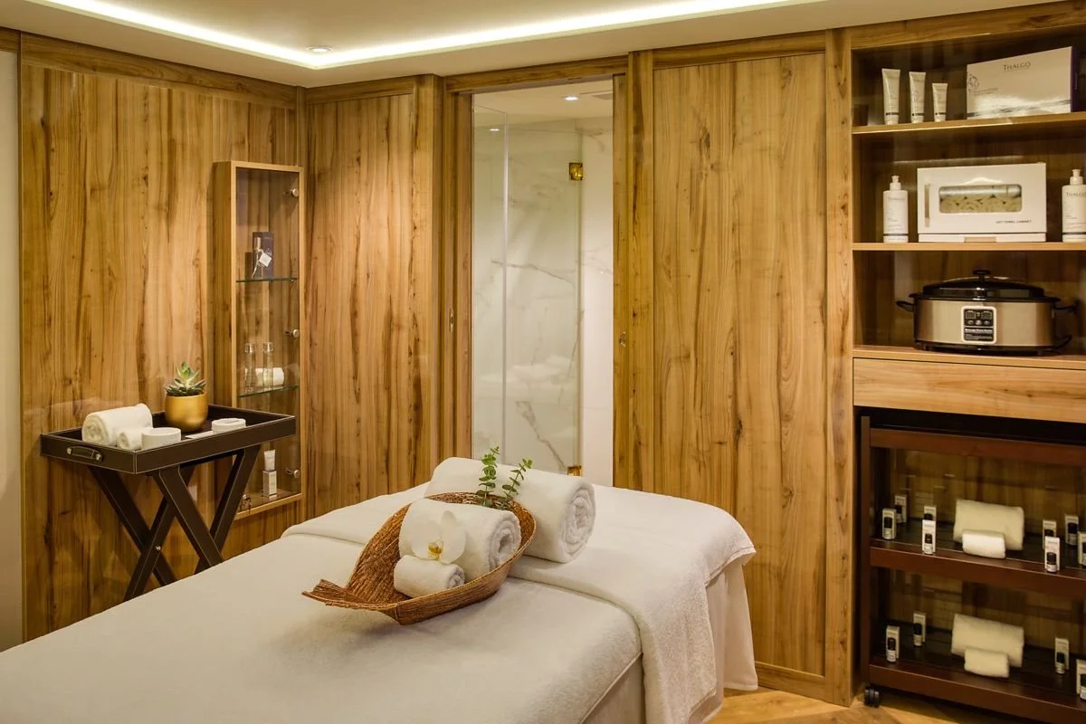 Spa treatment room with a massage bed covered in white towels, a wooden wall, a small tray with rolled towels and a plant, and shelves with skincare products and a slow cooker.