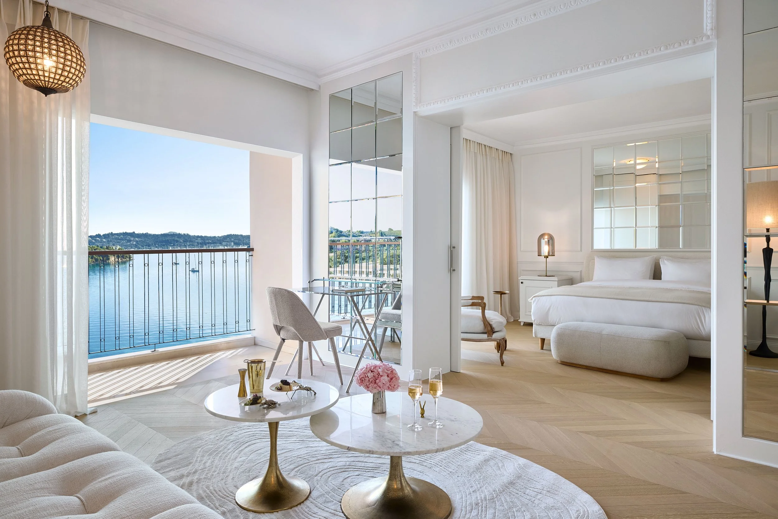 A bright living room with a large open window overlooking a body of water and hills. There is a white sofa, a round coffee table with a bouquet of pink flowers, and a dining area with chairs near the window. A bedroom with a white bed and a mirror-covered wall is visible through an open doorway. Elegant crown molding and white curtains are also present.