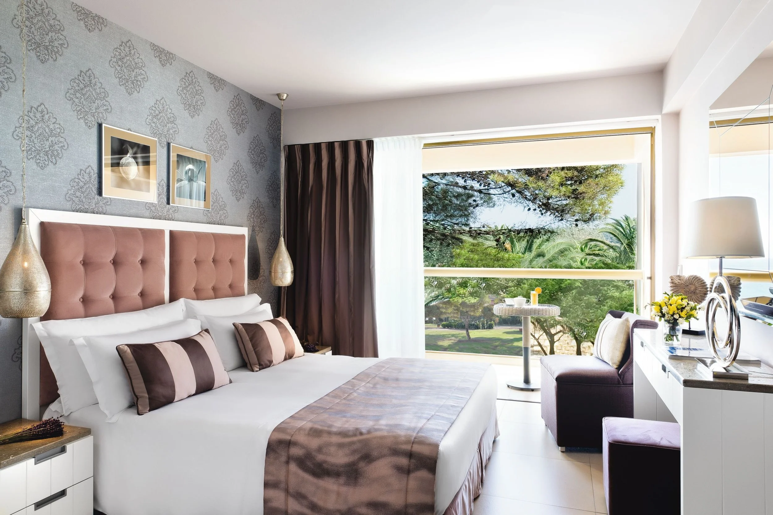 A hotel room with a bed, decorative pillows, and a large window showing green trees outside.
