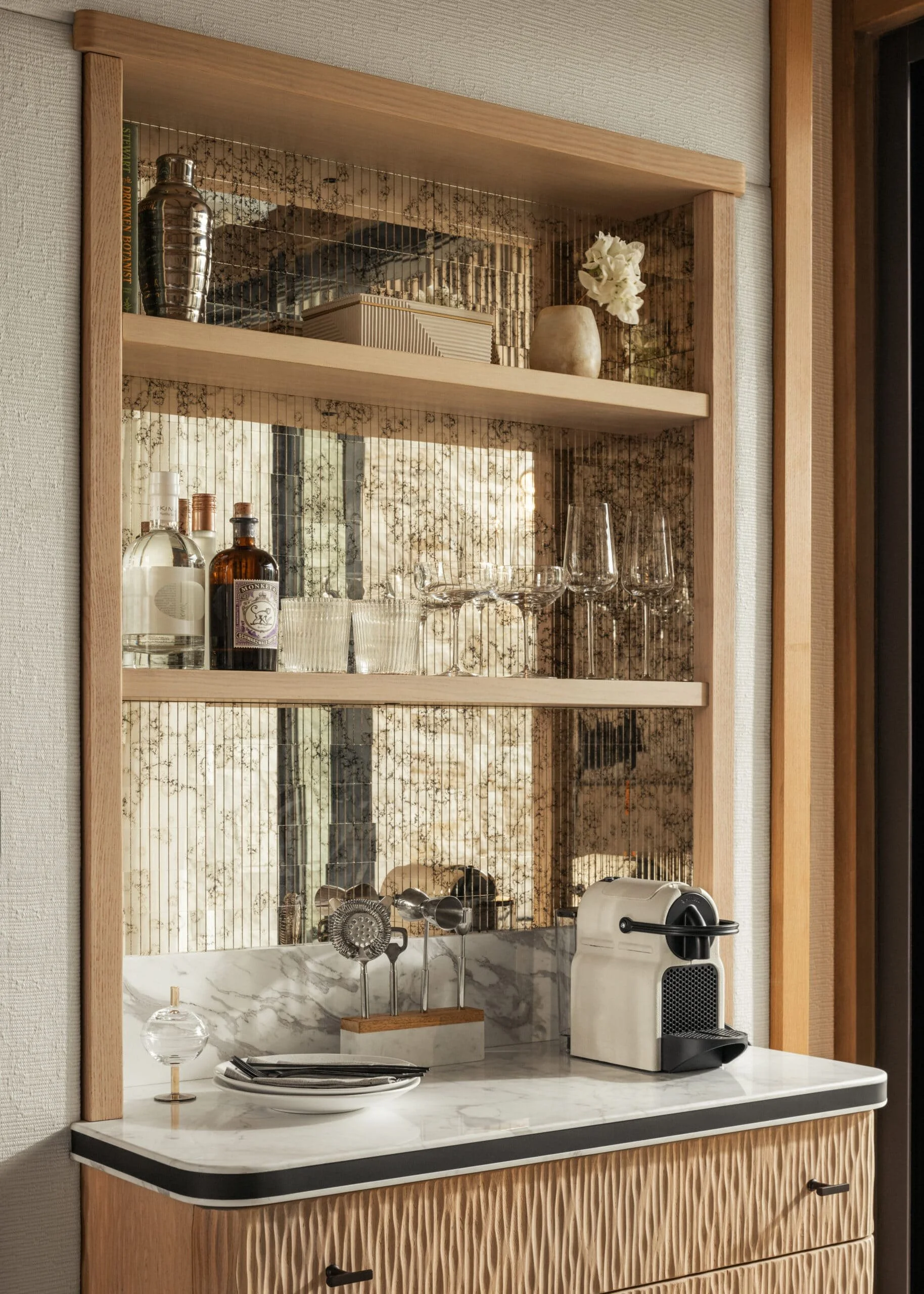 A small bar or kitchen area with a wooden frame, marble countertop, and decorative backsplash. Items on the shelves include bottles, glasses, and a flower vase. There is a coffee or espresso machine and some kitchen utensils on the counter.