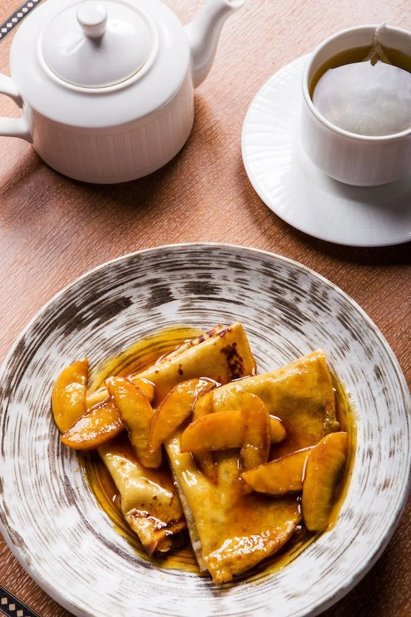 A bowl of chopped, cooked peaches in syrup, with a slice of pancake or crepe on top, on a decorative ceramic plate. In the background, a white teapot and a cup of hot tea on a saucer, sitting on a pink wooden table.
