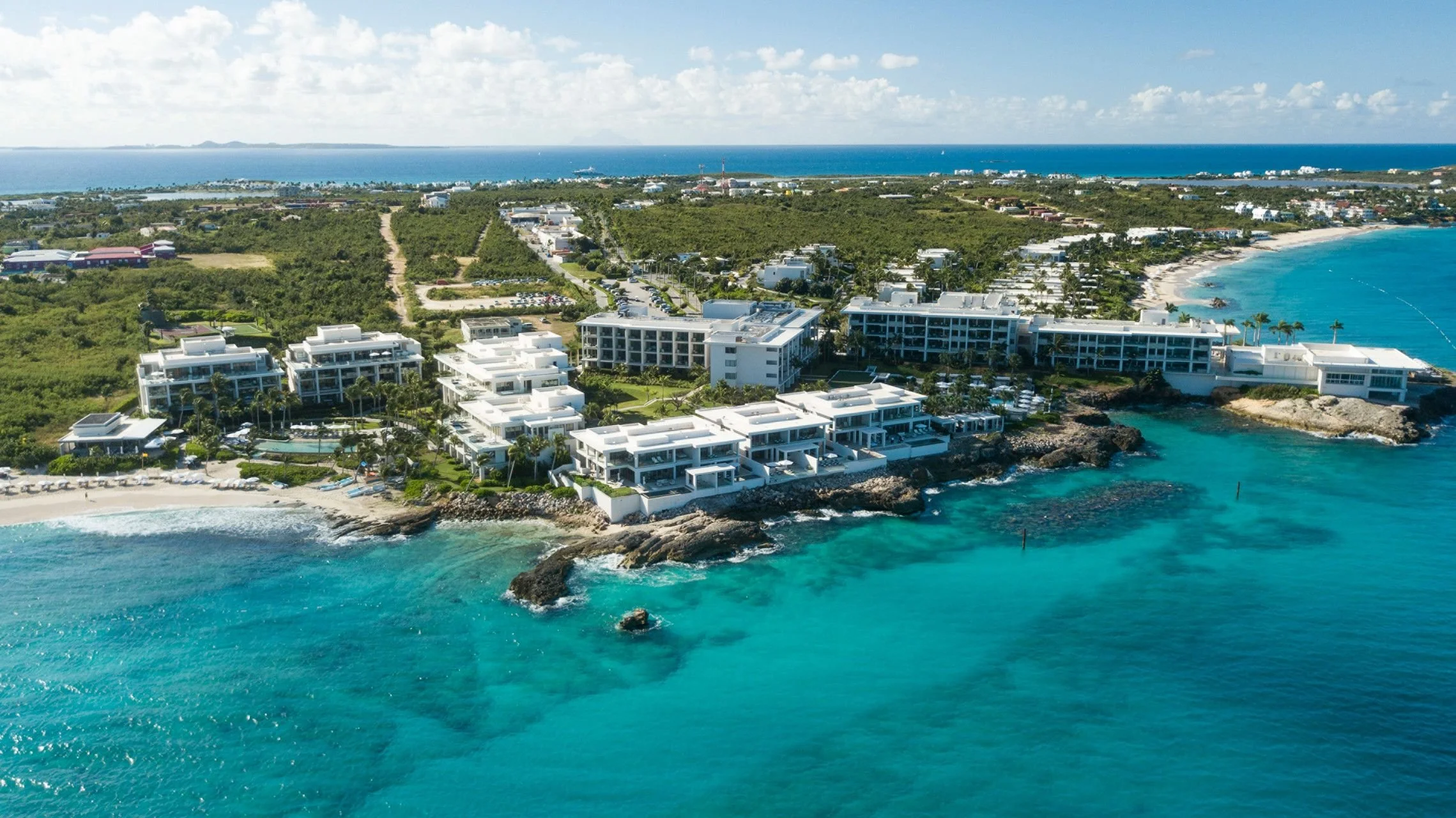 Aerial view of a beachfront resort with white modern buildings, palm trees, a sandy beach, and clear turquoise water, with a lush green landscape and distant ocean in the background.