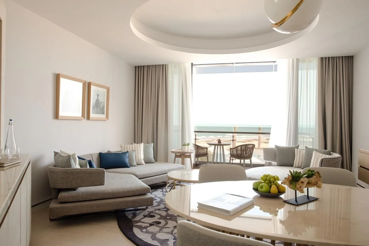 Modern living room with light-colored sofas, a round dining table with fruit centerpiece, a balcony with seating, large windows with curtains, and artwork on the wall.