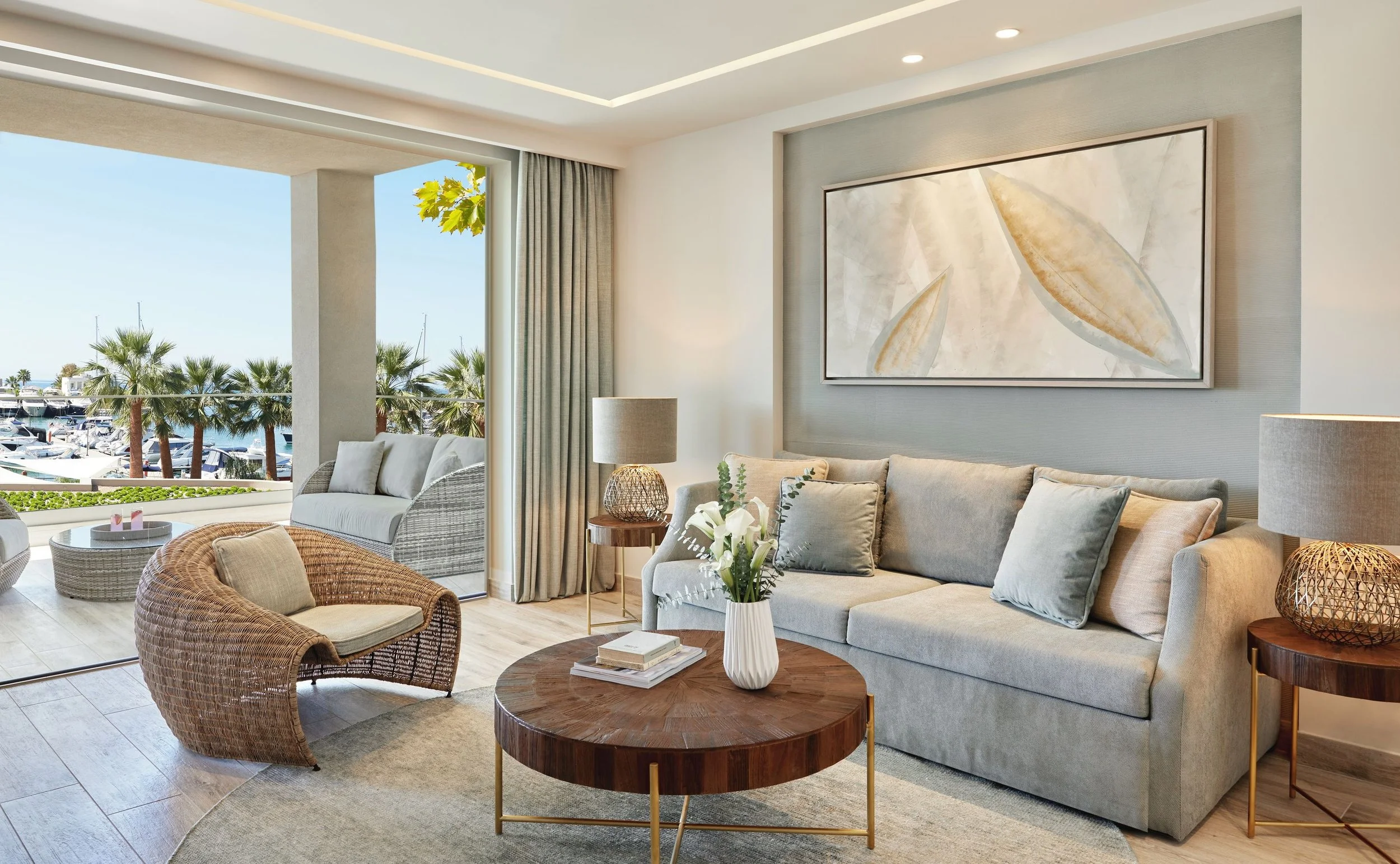 Living room with beige sofa, wicker chairs, coffee tables, and large window overlooking a marina with palm trees.