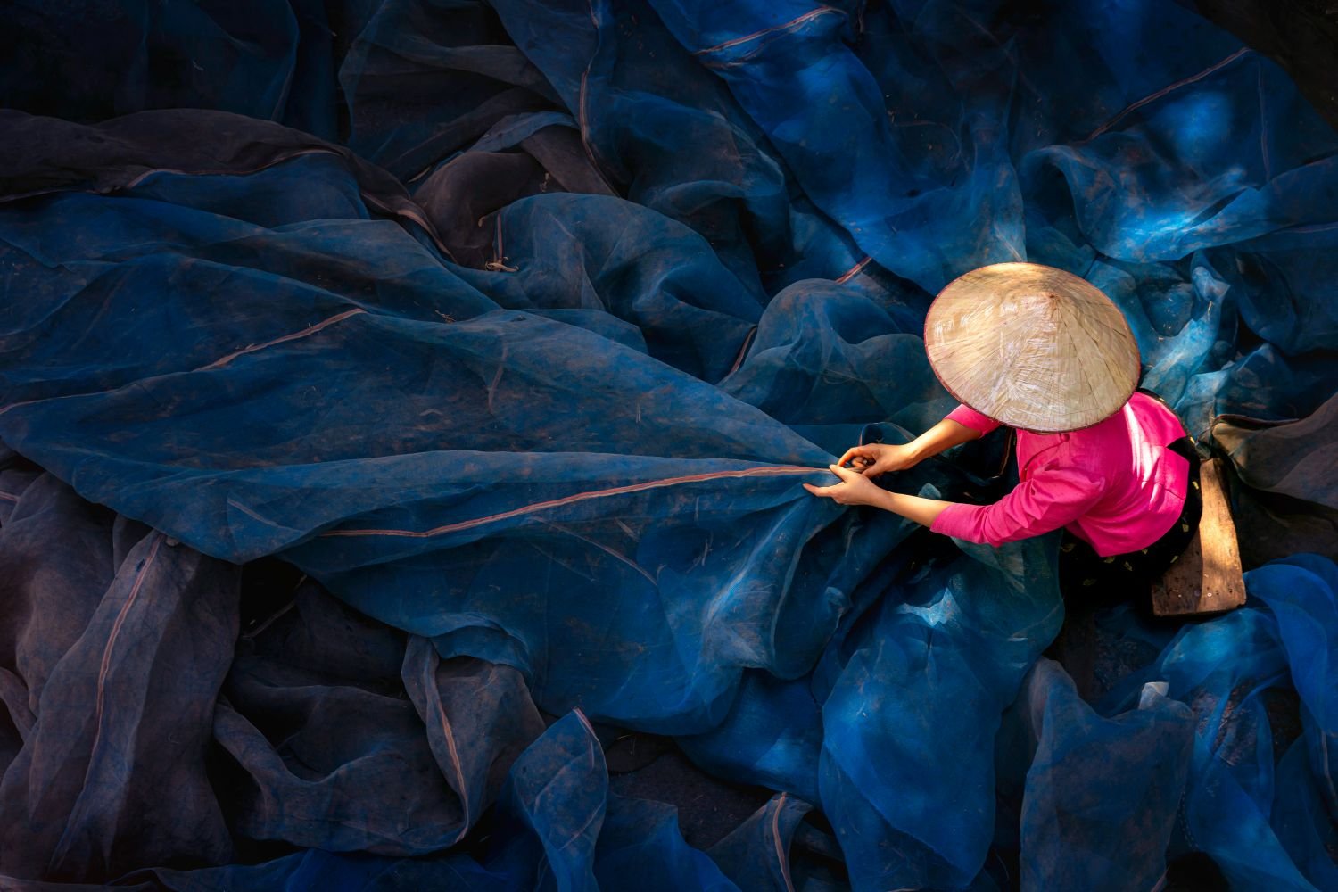 A person wearing a traditional conical hat and pink shirt is working with large blue plastic sheets.