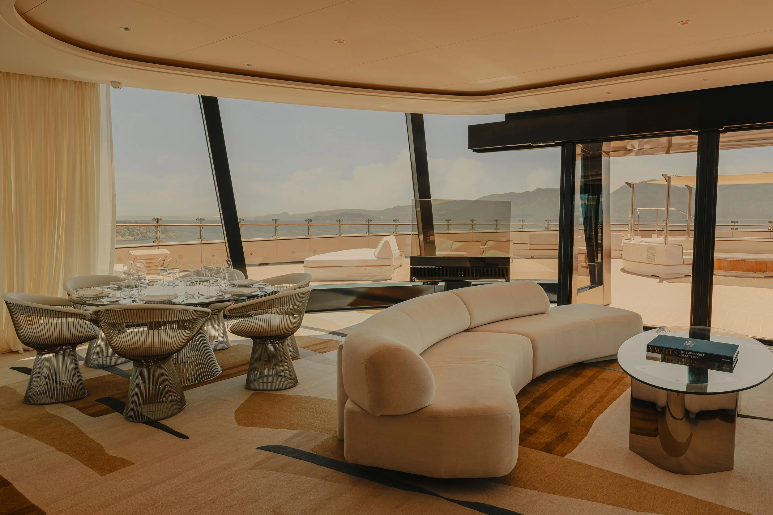 Living room with large glass windows showing an outdoor balcony and distant mountains, featuring a dining table set, a white curved sofa, a white round coffee table with books, and modern decor.