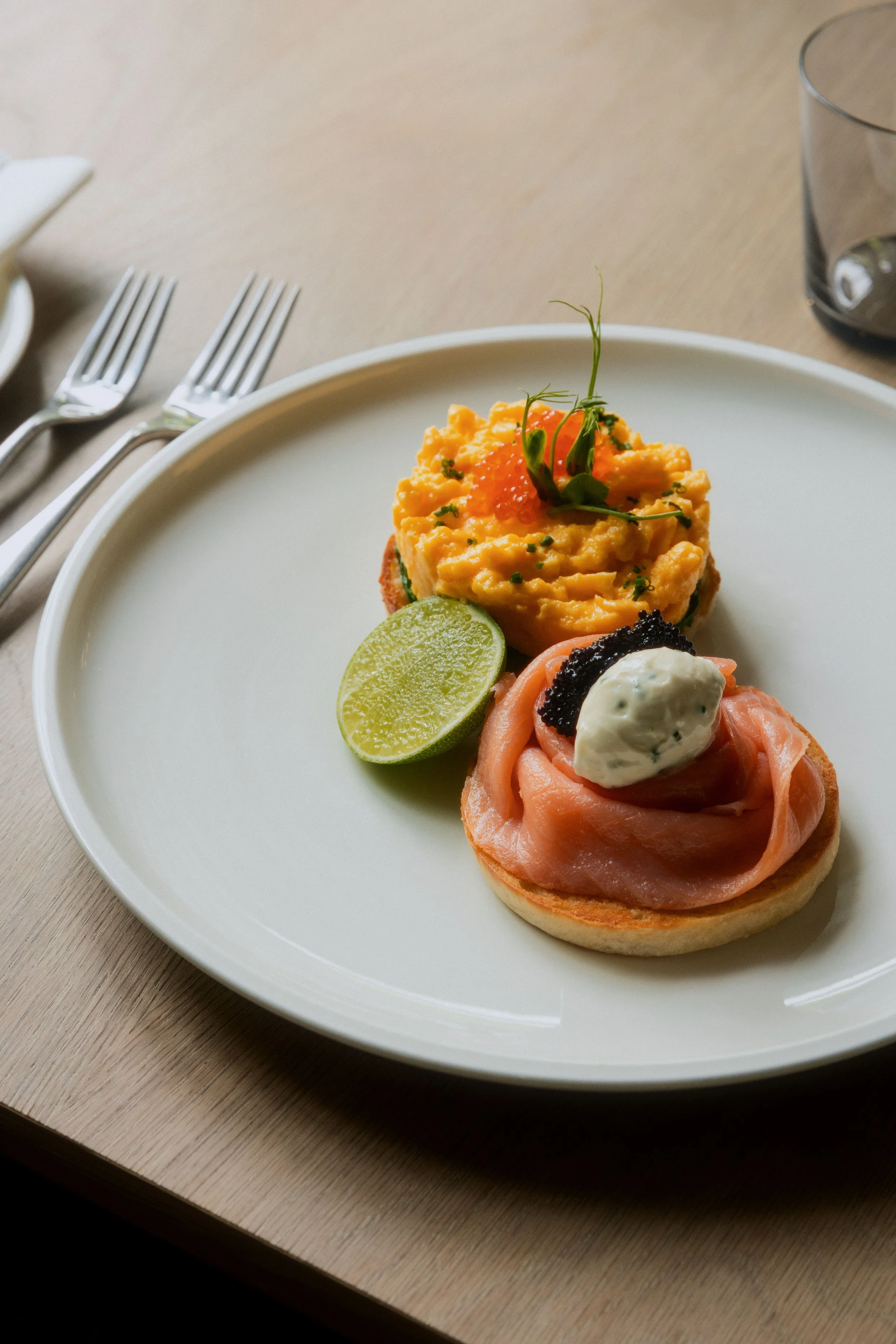 A white plate with two gourmet appetizers on a wooden table. One is topped with scrambled eggs, salmon, caviar, and microgreens. The other has smoked salmon, a dollop of herb cream, caviar, and lime wedge. Forks are to the side, and a glass of water is in the background.