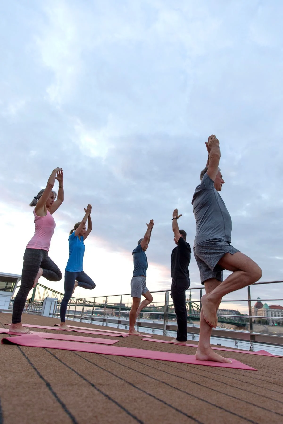 People practicing yoga outdoors on pink mats near a river, practicing tree pose with arms raised, with a cityscape and bridge in the background during cloudy weather.