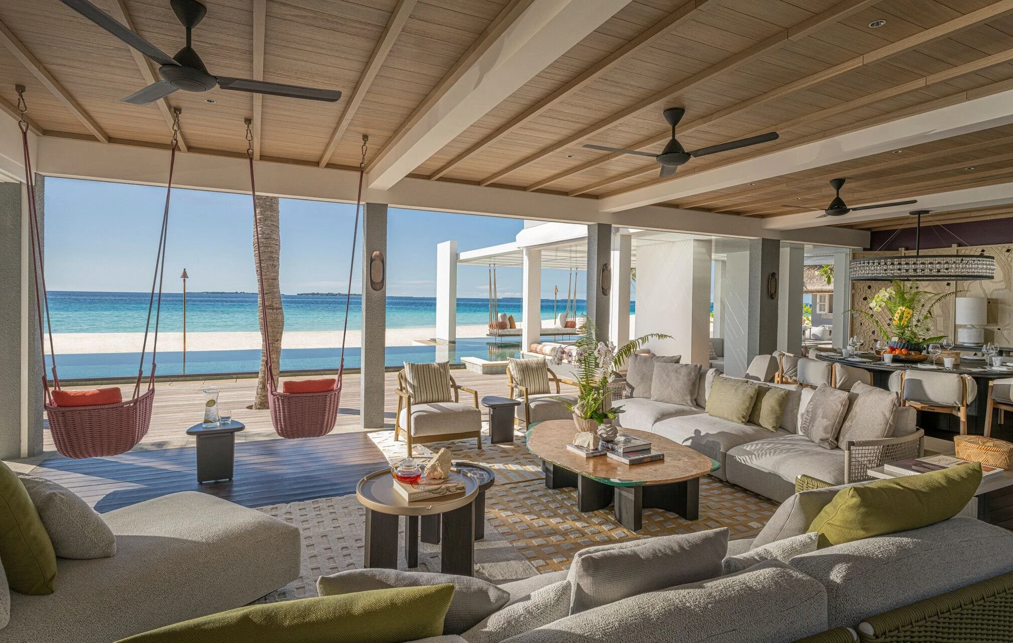 Living room with couches, coffee tables, and pillows, opening to a patio with hanging swings, overlooking a beach and ocean.