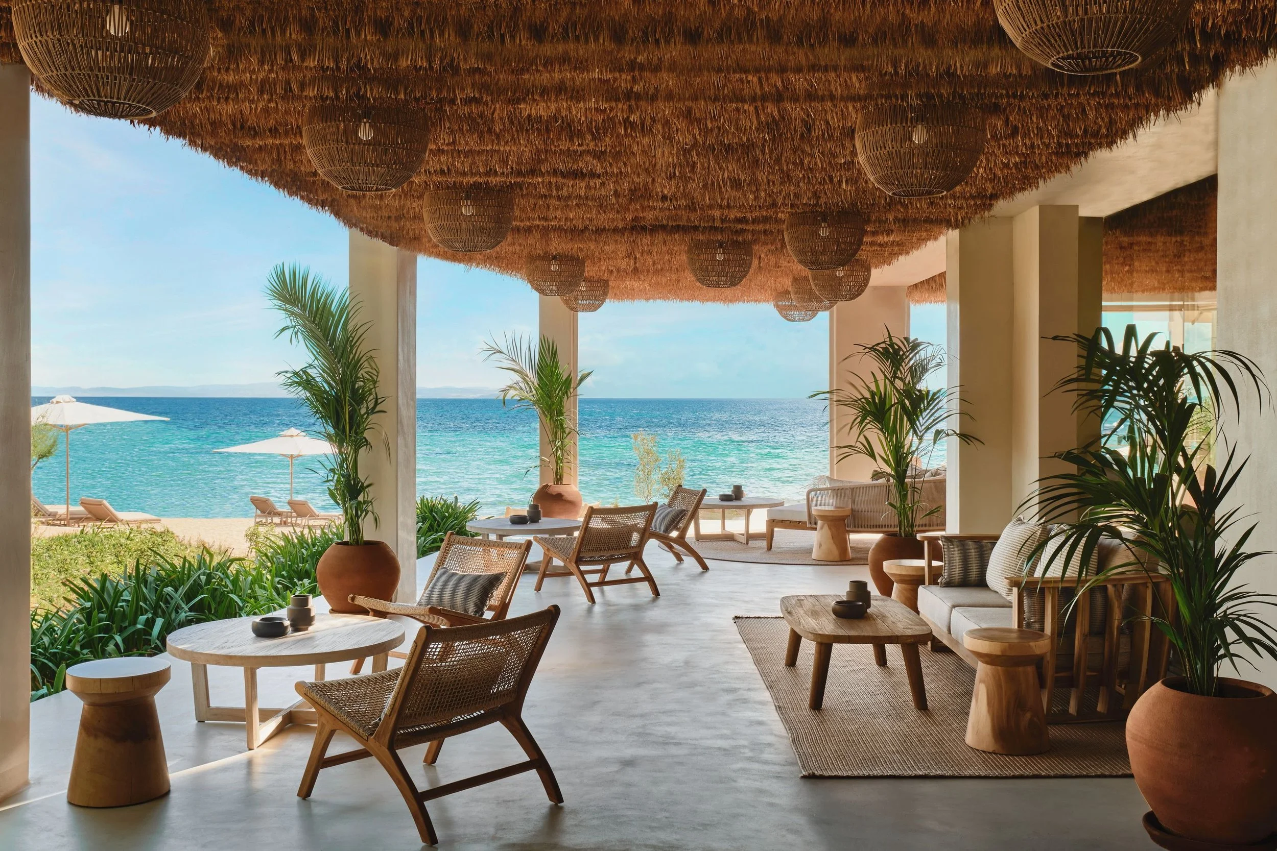 Open-air seaside restaurant with wooden furniture, potted plants, and a thatched roof, overlooking the ocean with beach umbrellas and chairs in the background.