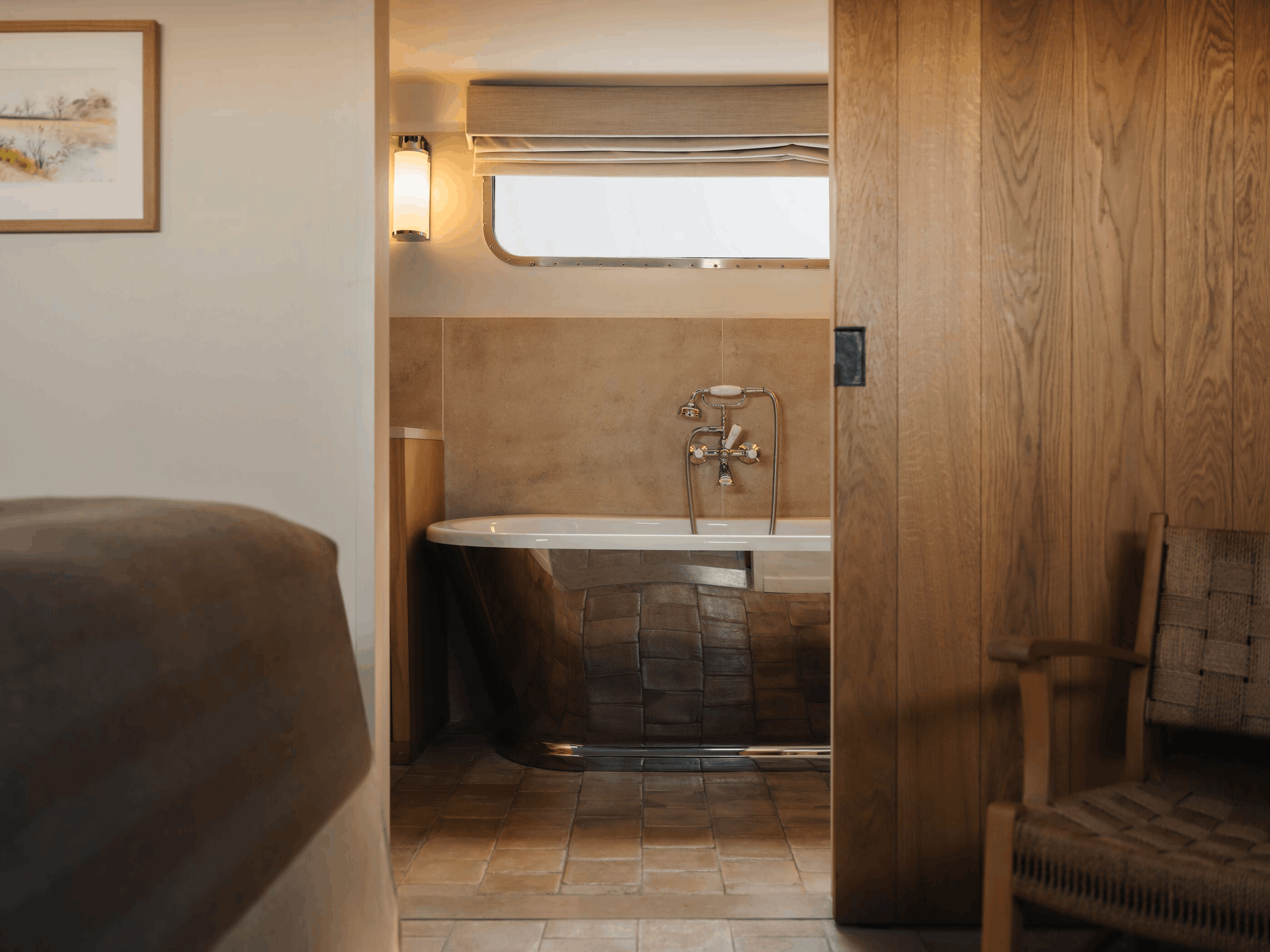 Interior view of a bathroom with a bathtub, a window with a beige blind, a wall-mounted light, and a wooden sliding door.