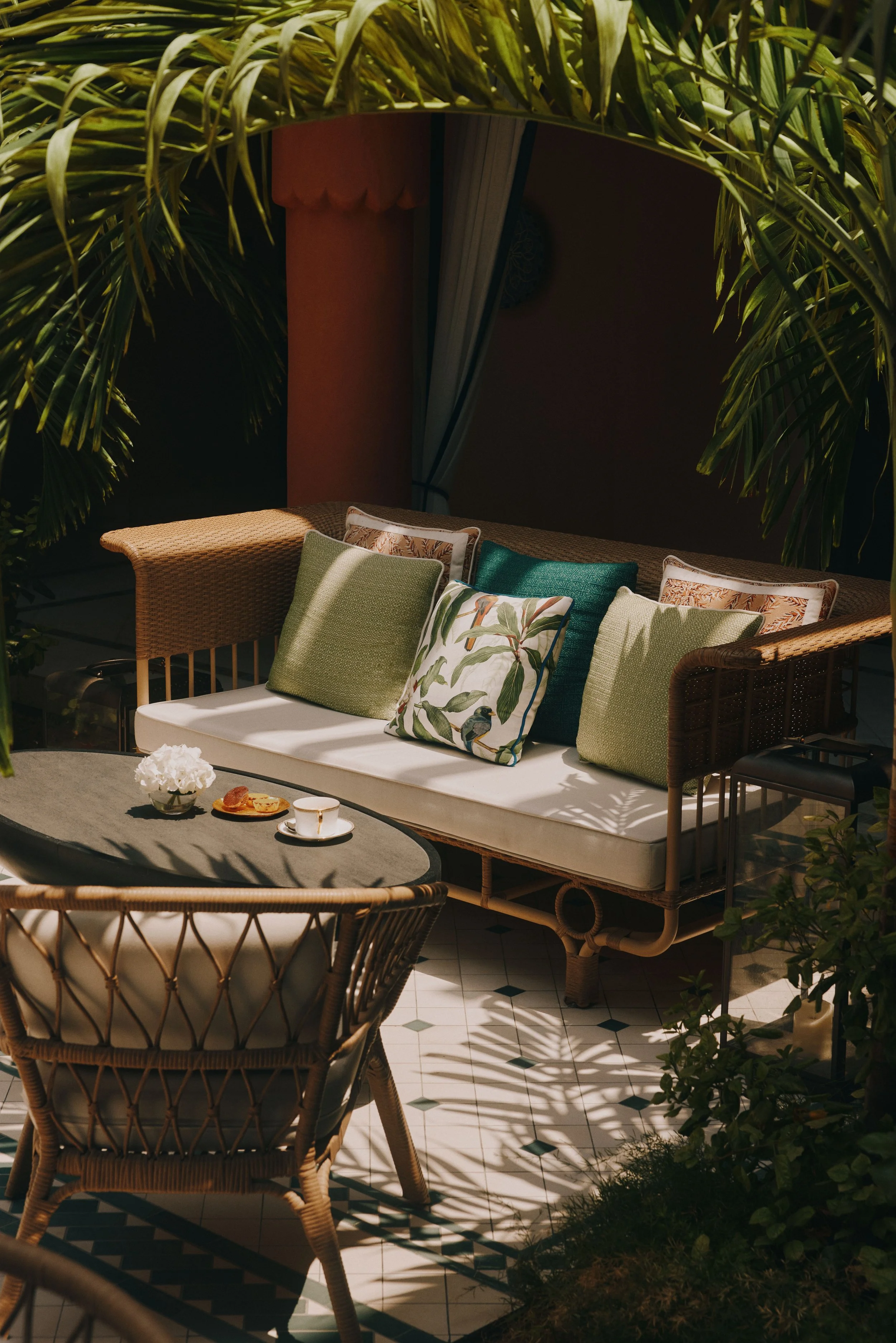 Outdoor patio with wicker sofa and chairs, colorful pillows, a coffee table with a flower arrangement and snacks, surrounded by lush green plants, under a covered canopy