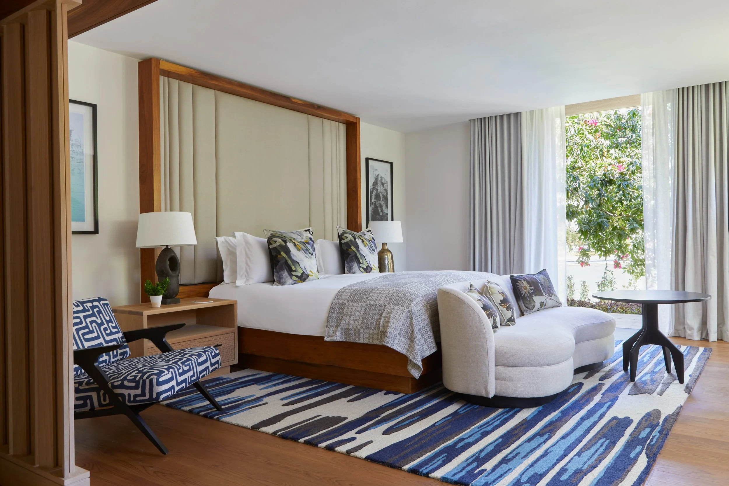 A bedroom with a large bed, decorative pillows, and white curtains beside a large window with greenery outside. There are lamps, framed pictures, a blue patterned chair, and a striped rug.