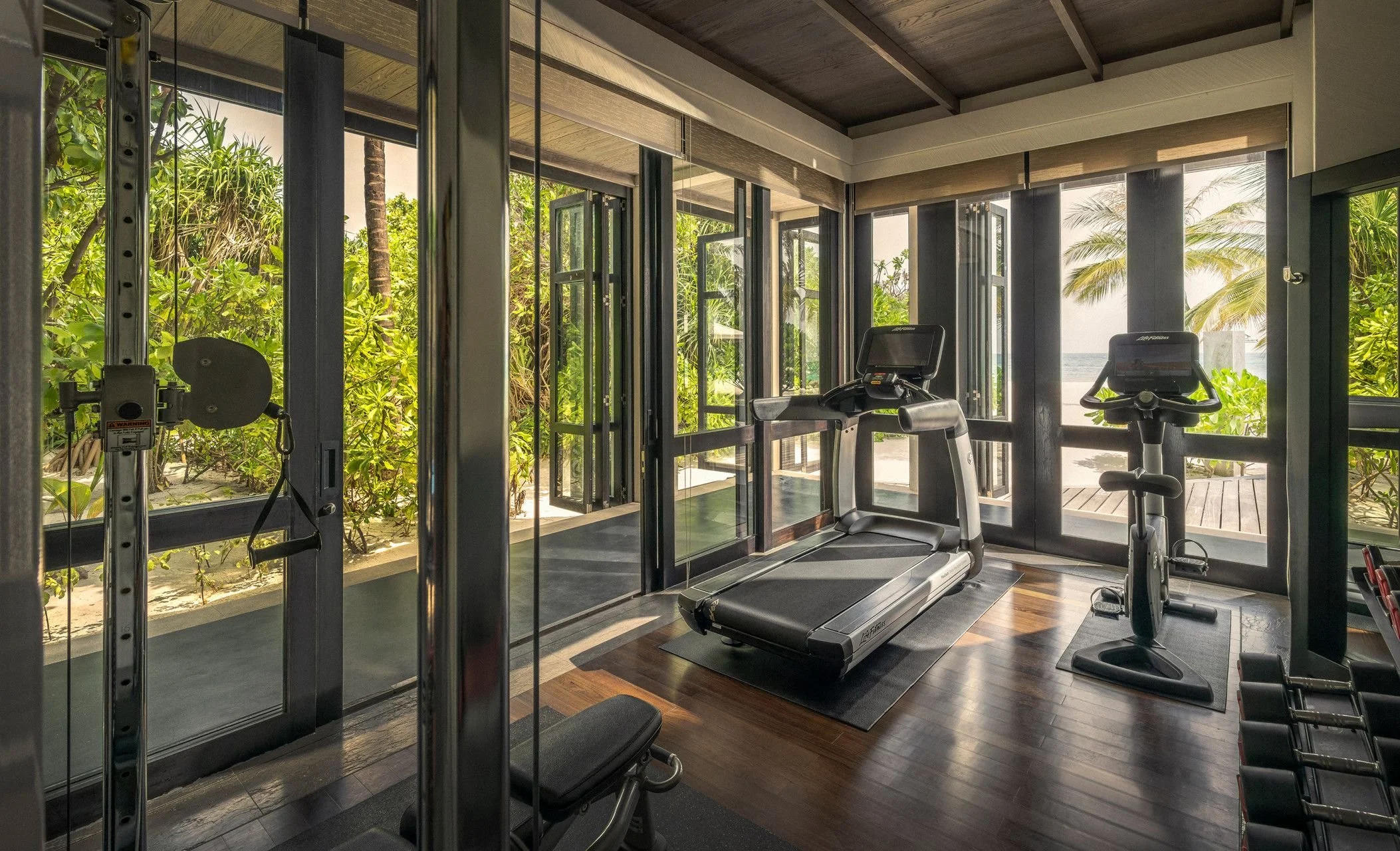 A private gym with large glass doors and windows showing tropical trees and a beach view outside. The gym has a treadmill, stationary bike, and weight equipment.