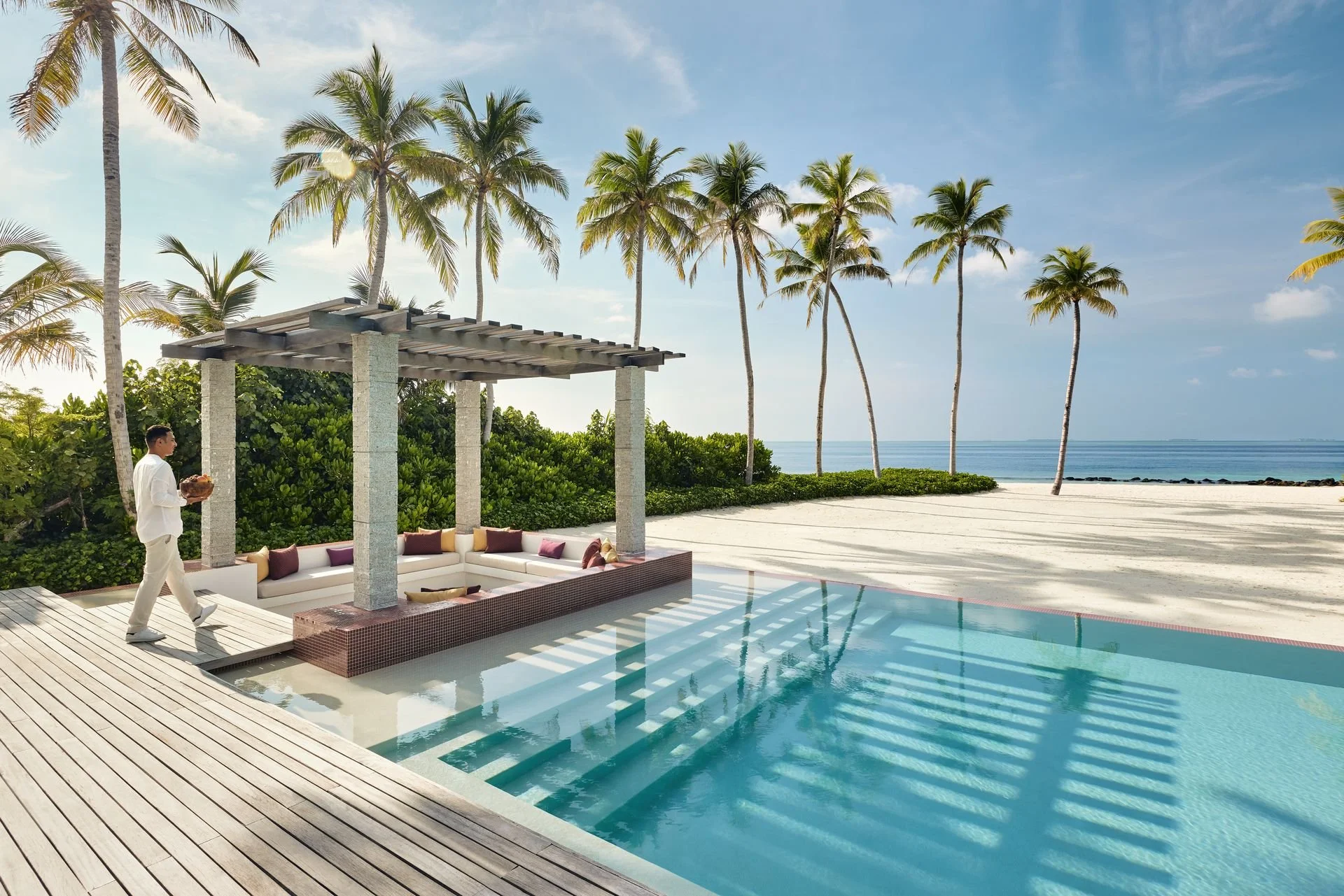 A luxury beachfront resort with a pool, tropical trees, and a sandy beach with the ocean in the background. A person dressed in white walking near a shaded lounge area.