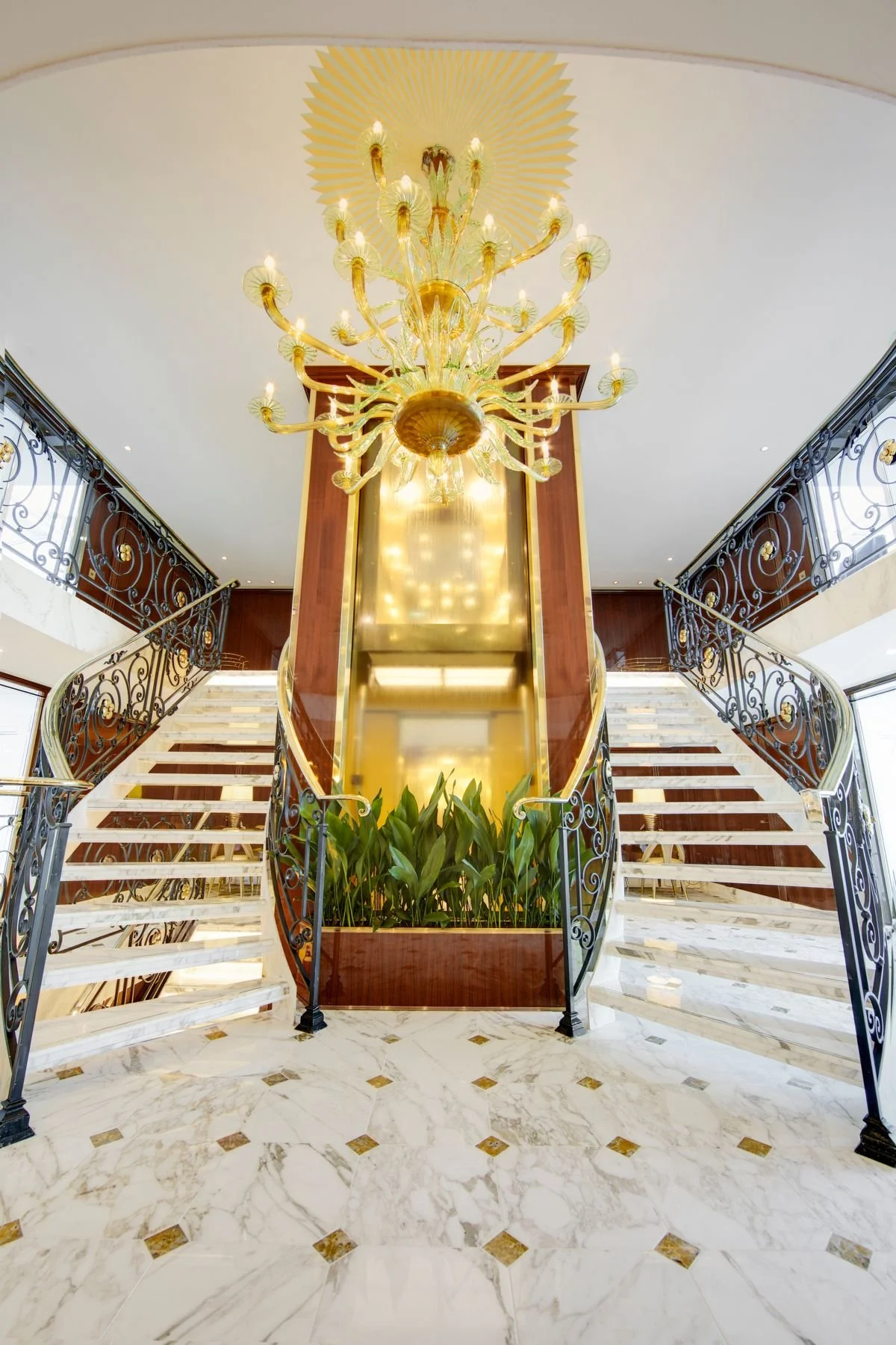 Luxurious hotel lobby with a grand staircase, marble flooring, ornate wrought iron railings, a large gold chandelier, and plants at the base of the staircase.
