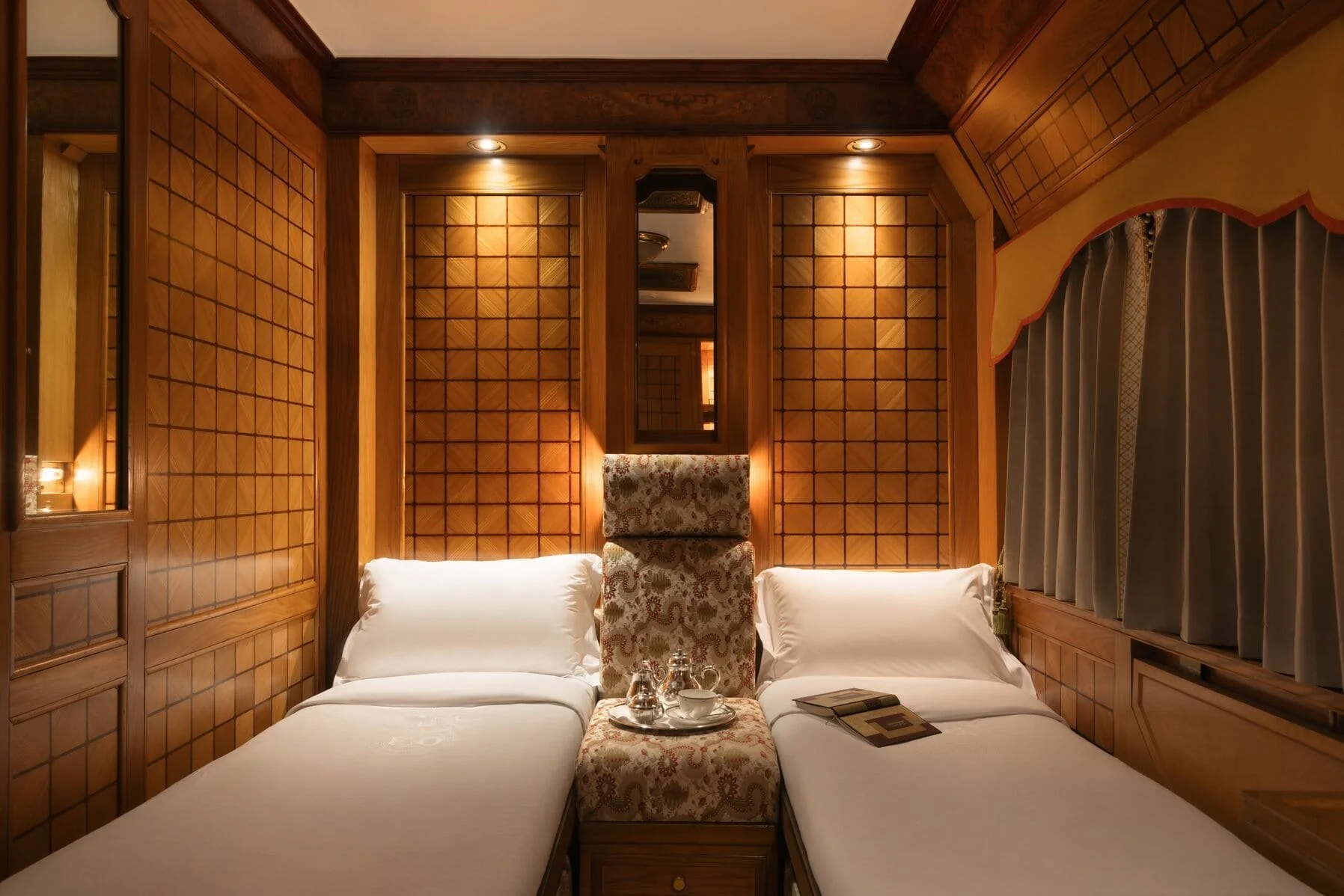 A cozy hotel room with two twin beds, a central high back upholstered headboard, and wooden walls with a grid pattern. There is a mirror on the wall, bedside lamps, and a window with curtains.