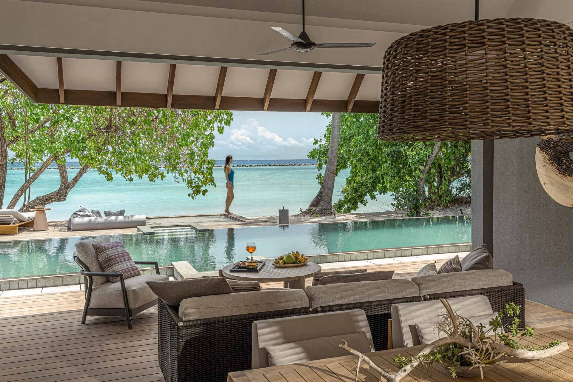 View from an indoor-outdoor living space overlooking the ocean. There is a woman in a blue swimsuit standing on the deck near the water, and a swimming pool in the foreground. The area has greenery and trees, with outdoor furniture including chairs, a sofa, and a table with a glass of orange drink and a bowl of fruit.