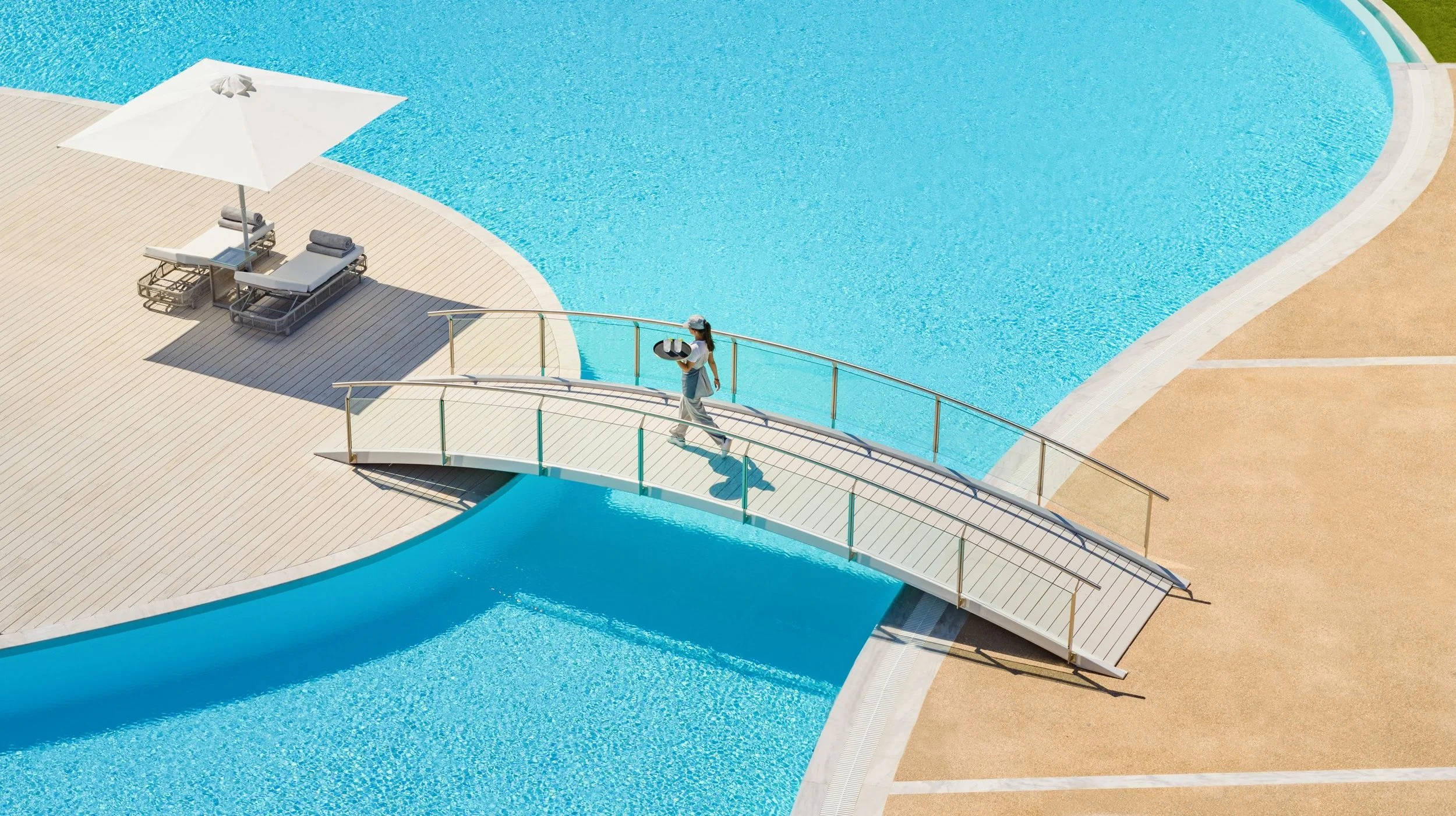 A spacious outdoor swimming pool with clear blue water, a curved white bridge, and lounge chairs with a white umbrella, on a bright and sunny day.