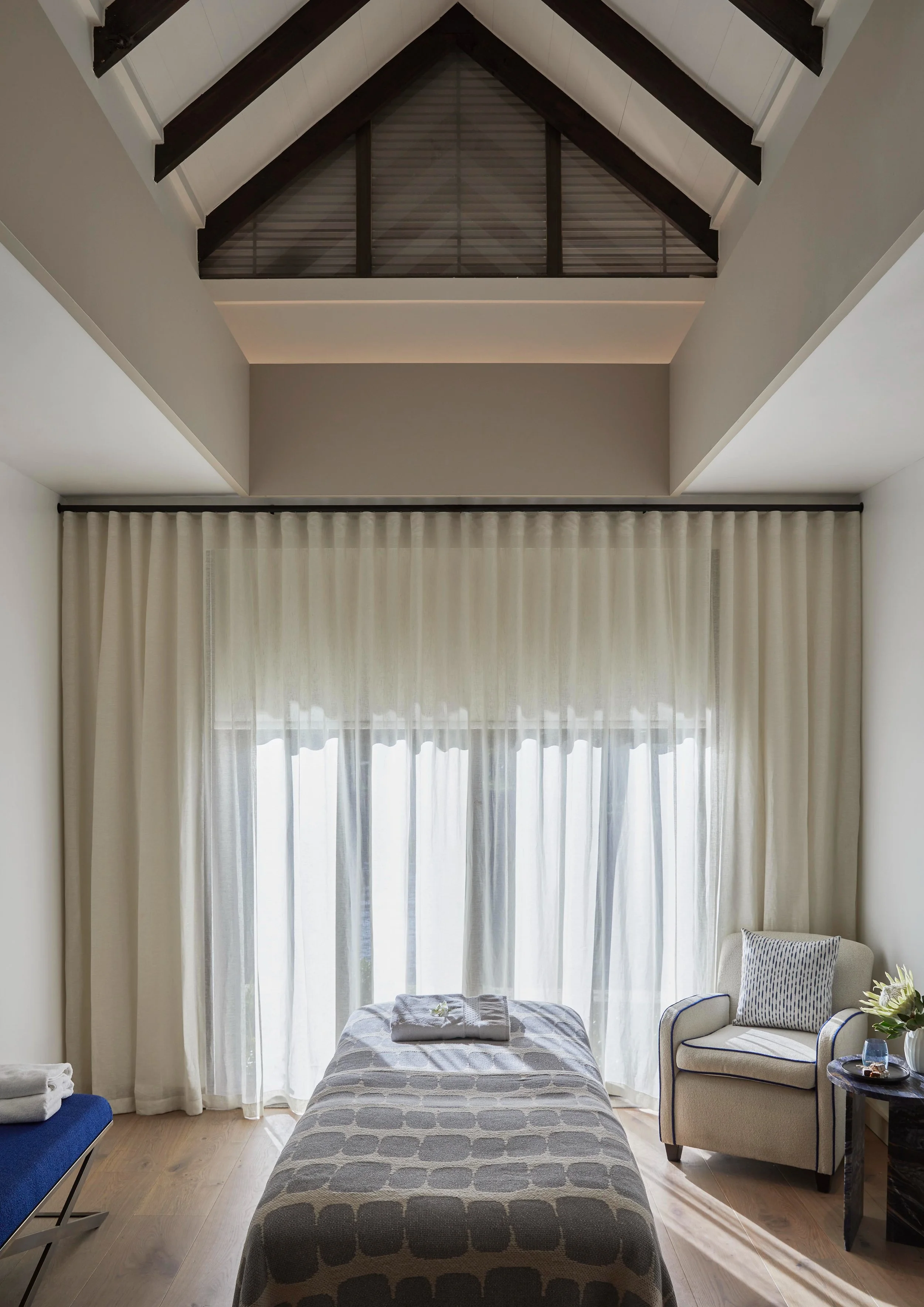 Interior of a bright room with a bed, a beige armchair with blue piping, a small side table with flowers, and large window with sheer curtains letting in natural light. The ceiling has exposed dark wooden beams.