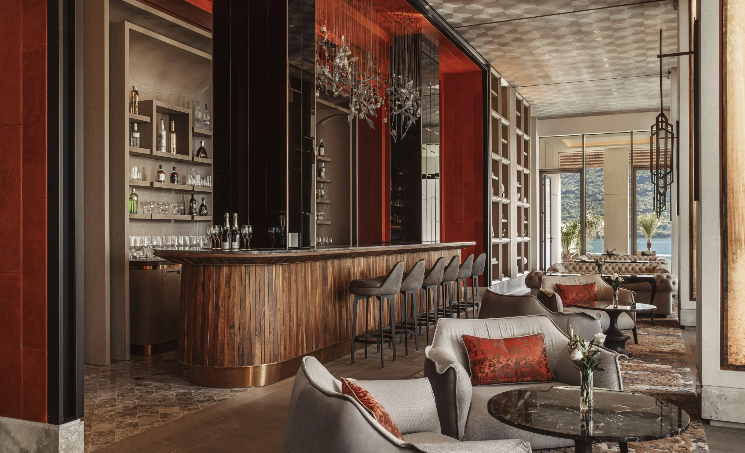 Interior of a luxurious lounge or hotel bar with a wooden counter, barstools, plush seating, large windows showing a water view, modern chandelier, and decorative shelving.