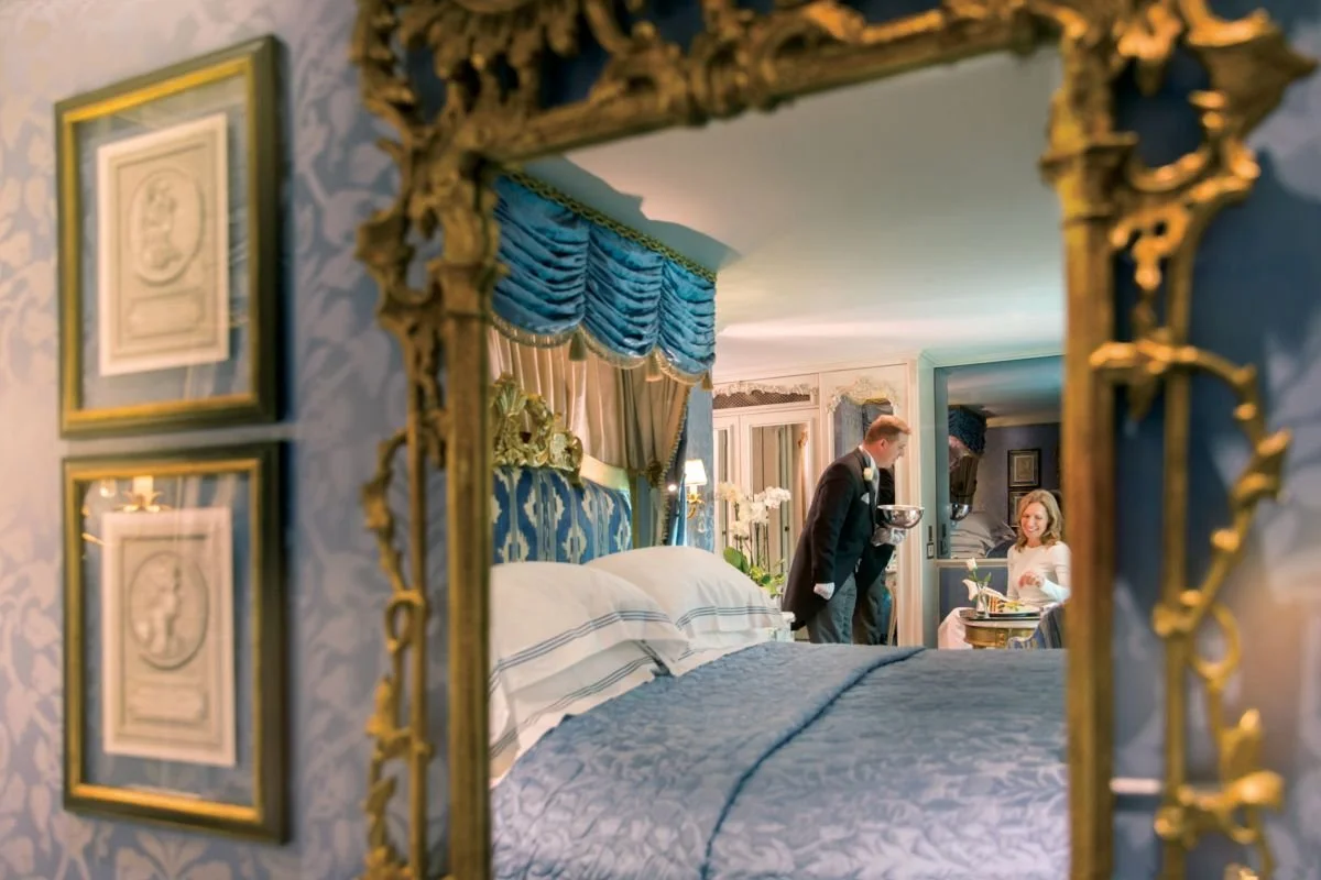 A reflection in an ornate gold-framed mirror shows a luxurious hotel room with a bed, blue curtains, and a woman sitting at a table, with a man in a suit serving her.