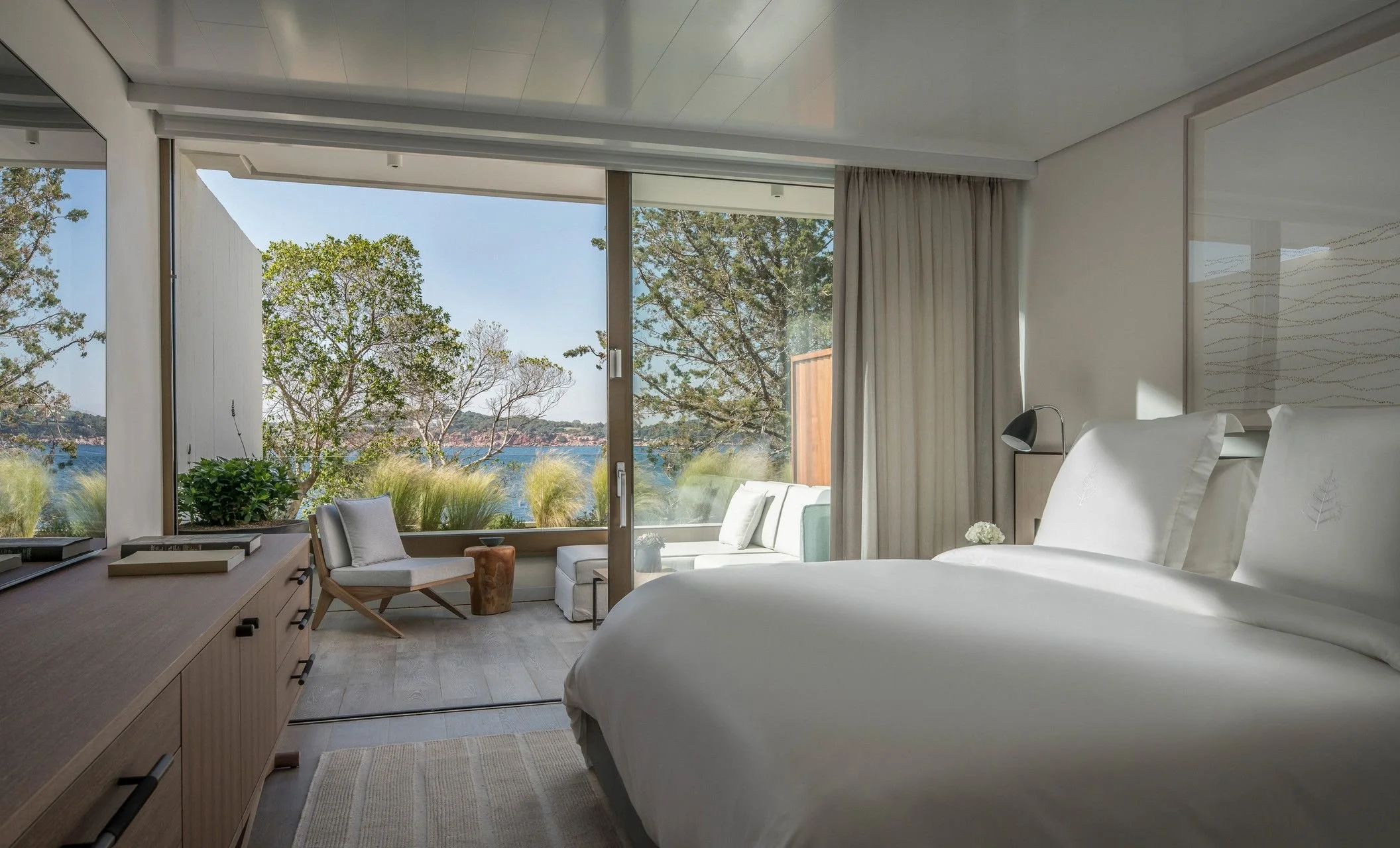 A modern bedroom with large floor-to-ceiling windows showing a view of trees and water outside. The room has a large bed with white linens, a bedside lamp, a small armchair, and a wooden dresser with books and decor.