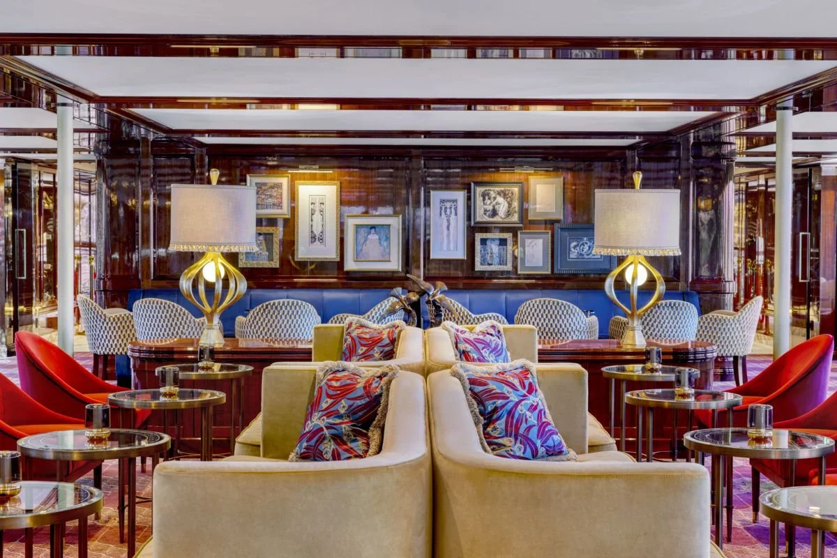 Elegant hotel lounge with plush beige armchairs, colorful pillows, marble side tables, decorative lamps, and framed artwork on dark wood-paneled walls.