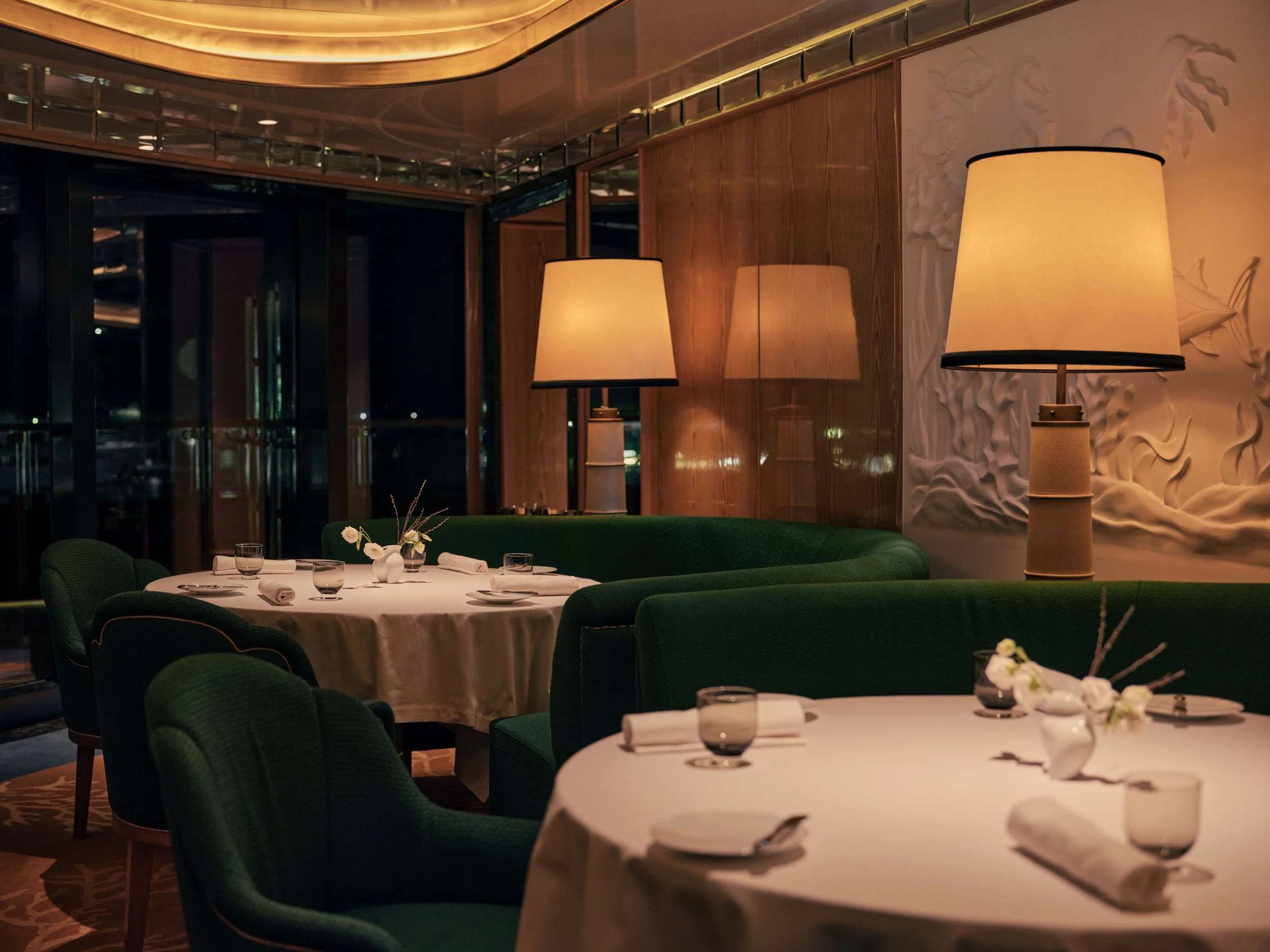 Elegant restaurant interior with round tables set with white tablecloths, glassware, napkins, and floral centerpieces. Green chairs surround the tables, and warm lighting is provided by tall lamps. Wooden and glass decor features are visible on the walls and ceiling.
